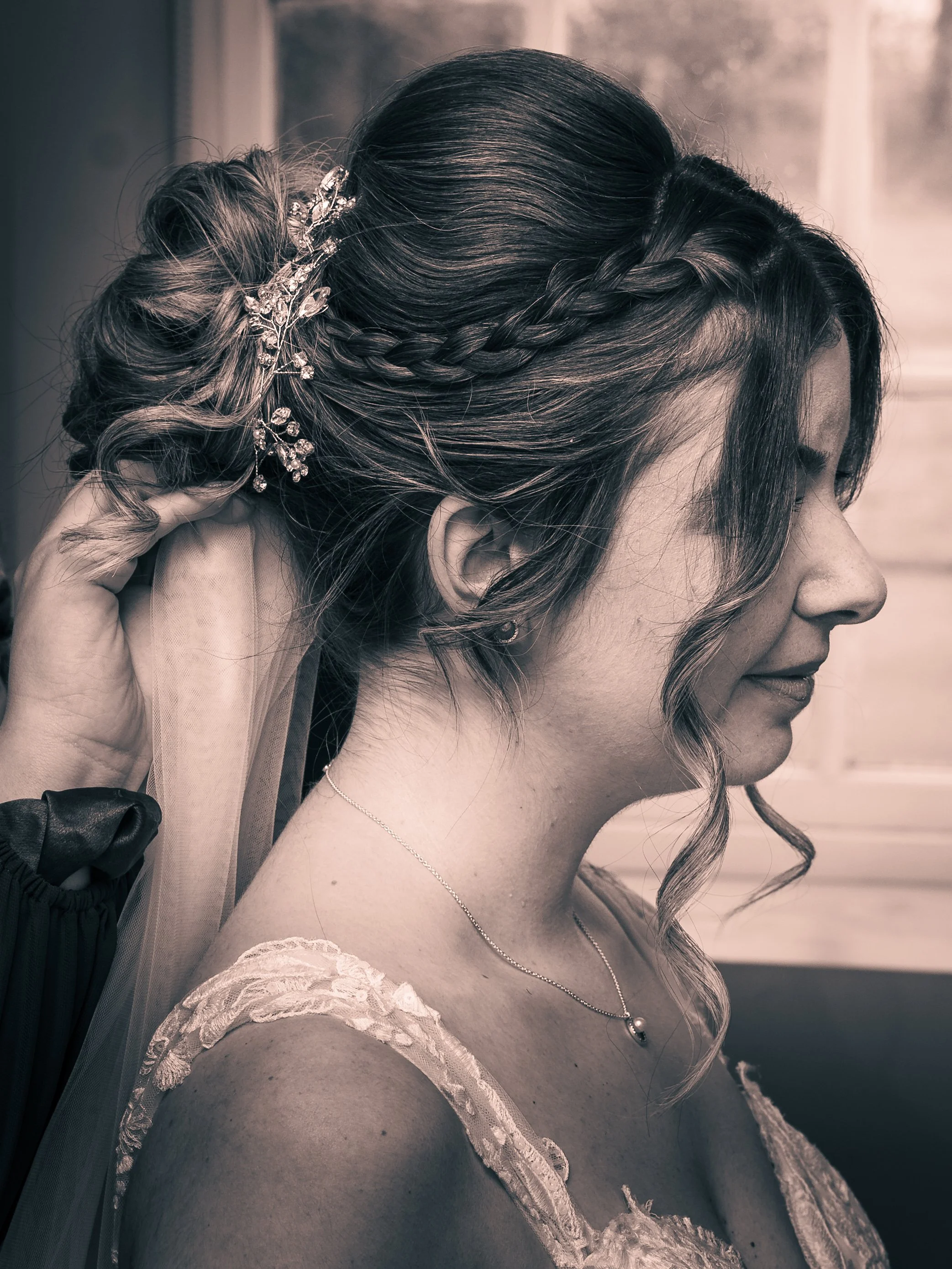 A woman with styled hair and a decorative hairpiece, wearing a lace dress and a necklace, is having her hair done.