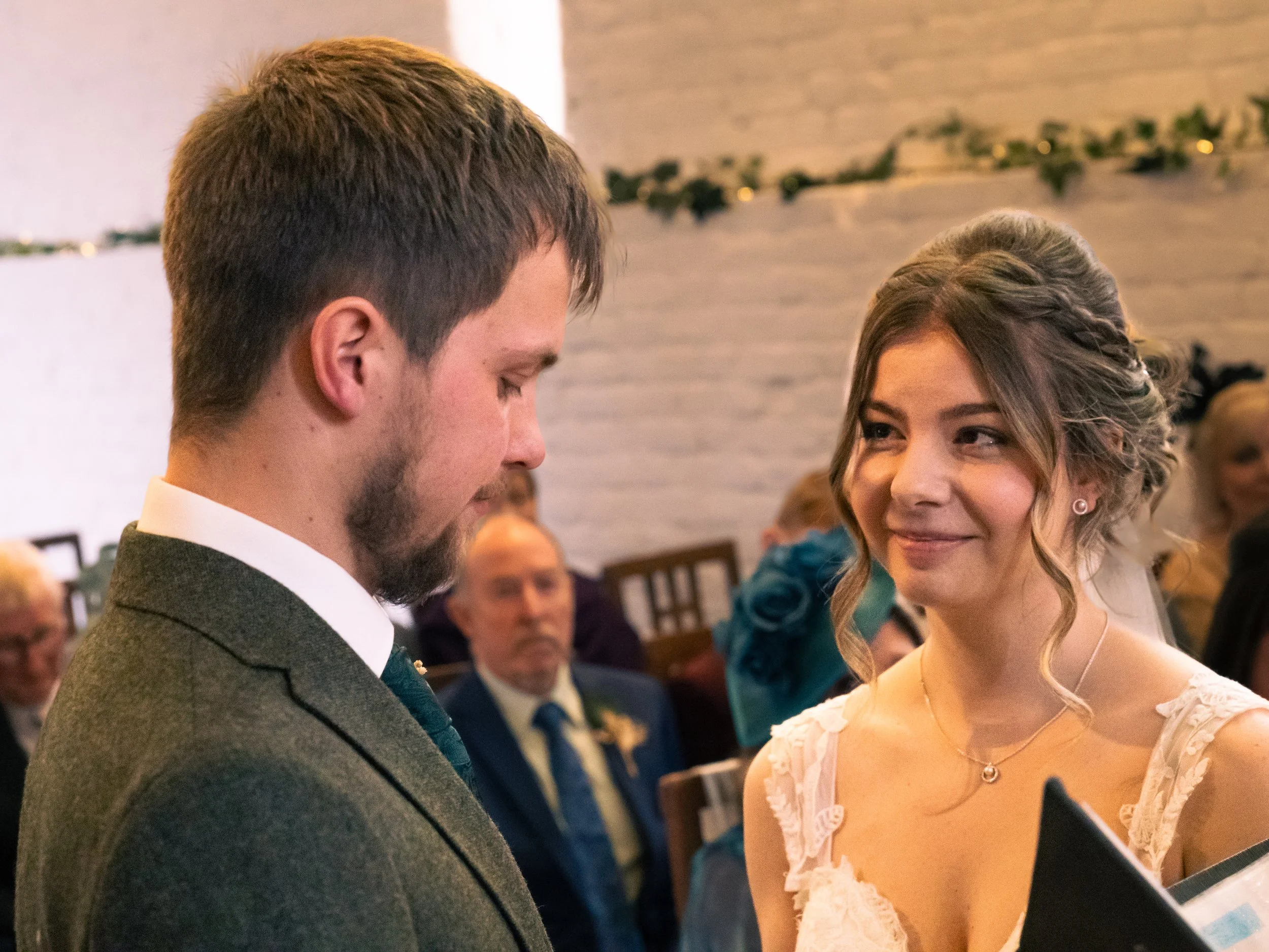 A groom and bride during a wedding ceremony. The groom has light brown hair, a beard, and is wearing a grey suit. The bride has long wavy hair, wearing a white dress with lace details, and a necklace. They are gazing at each other with a background o