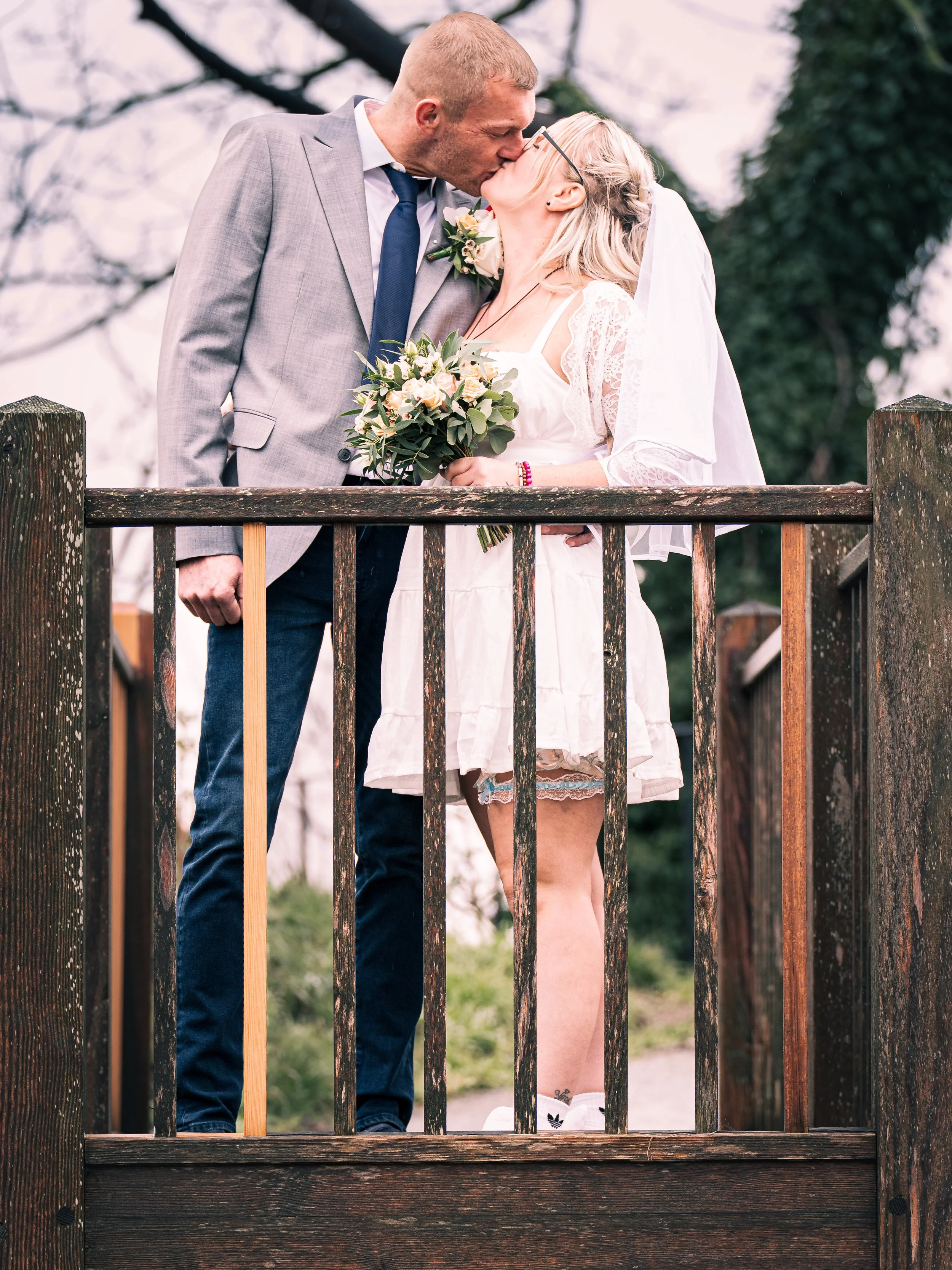 A couple on a bridge sharing a kiss during their wedding, the man in a gray suit and the woman in a white dress holding a bouquet of flowers.