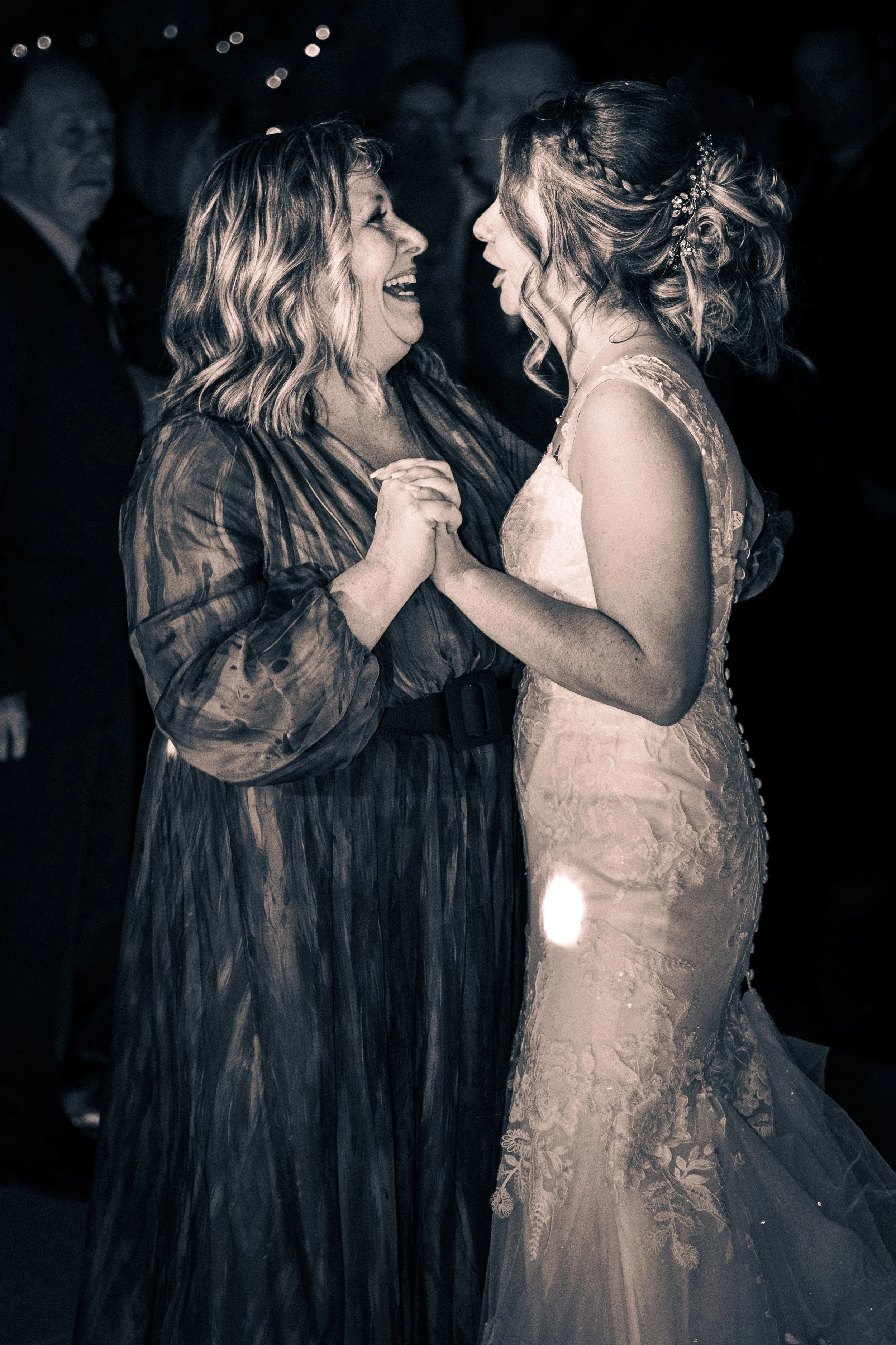 A woman in a striped dress dancing and holding hands with a woman in a wedding gown at a celebration, both smiling and appearing joyful.