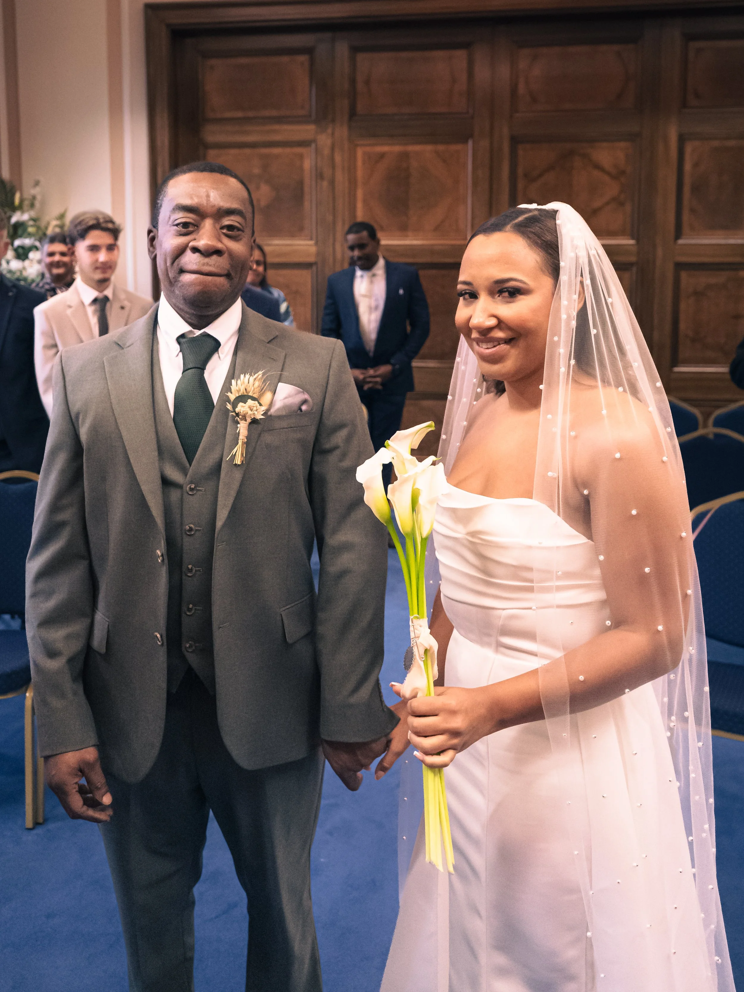 A bride and an older man holding hands at a wedding ceremony, with guests in the background, in a formal indoor setting.