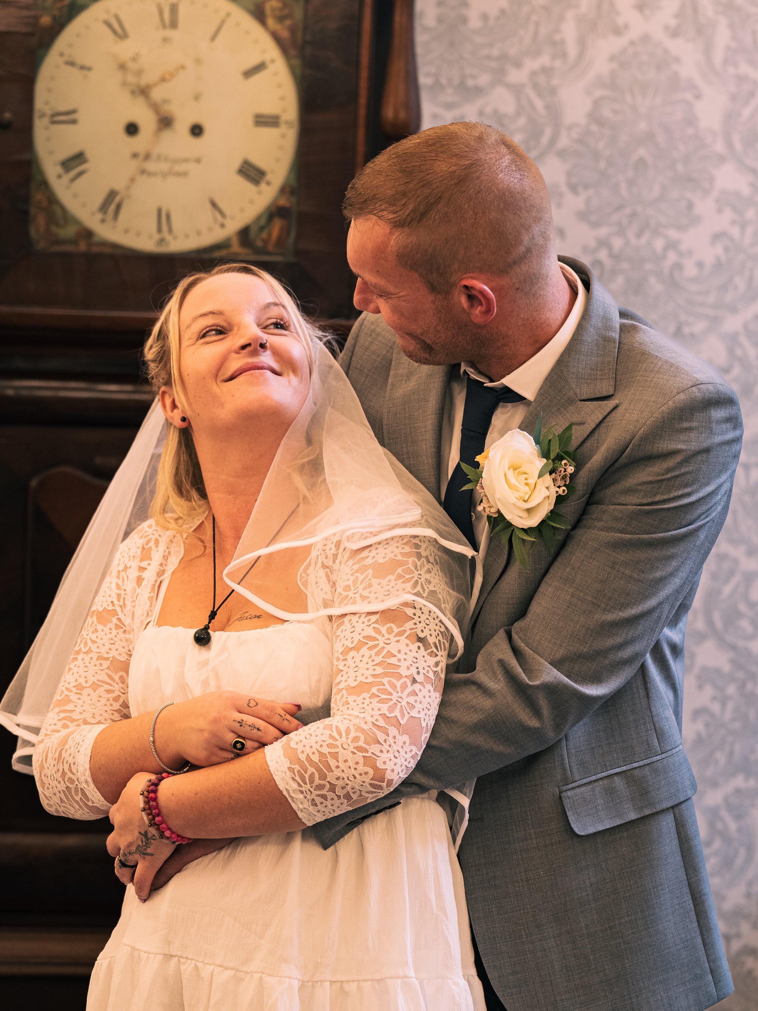 A bride and groom sharing a moment indoors, with the groom hugging the bride from behind. The bride is smiling and looking up at the groom. The bride is wearing a lace dress and veil, with jewelry and tattoos on her arms. The groom is wearing a gray 
