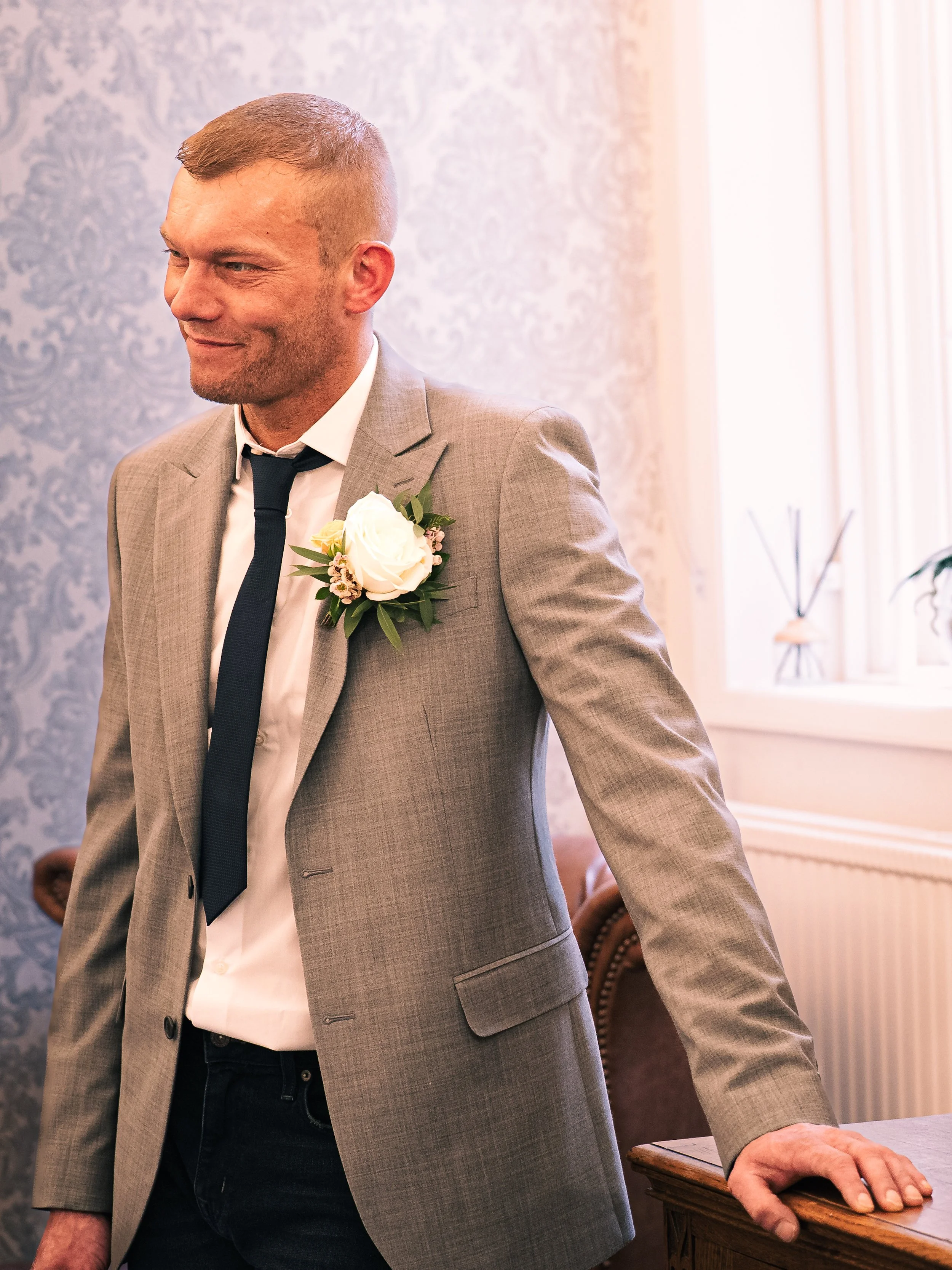 A man in a gray suit with a white shirt and dark tie, wearing a boutonniere with white flowers and greenery, standing in a room with patterned wallpaper and a window with sunlight, leaning on a wooden table.
