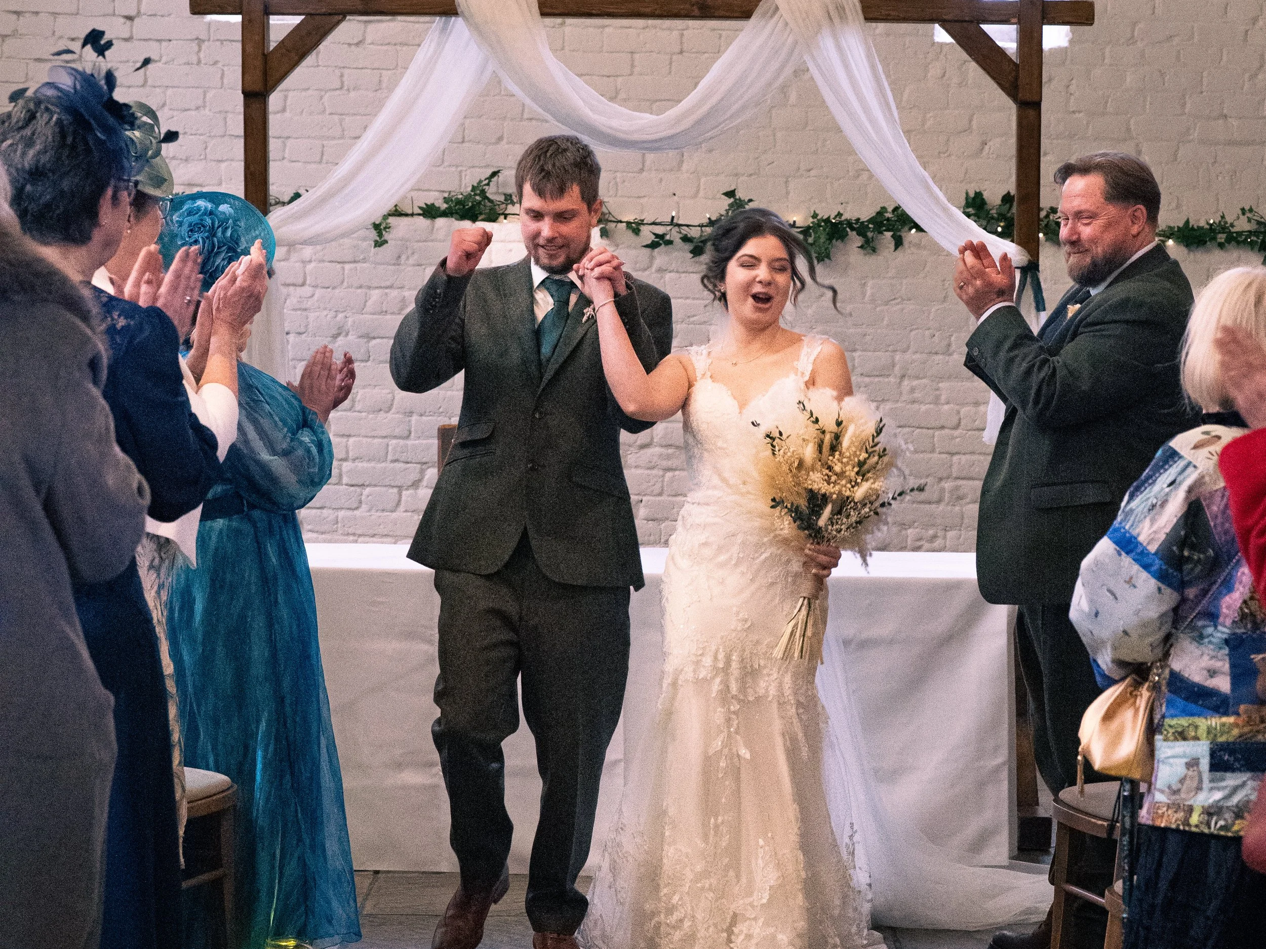 Bride and groom celebrating after wedding ceremony, holding hands and smiling, surrounded by applauding guests indoors with decorated backdrop.