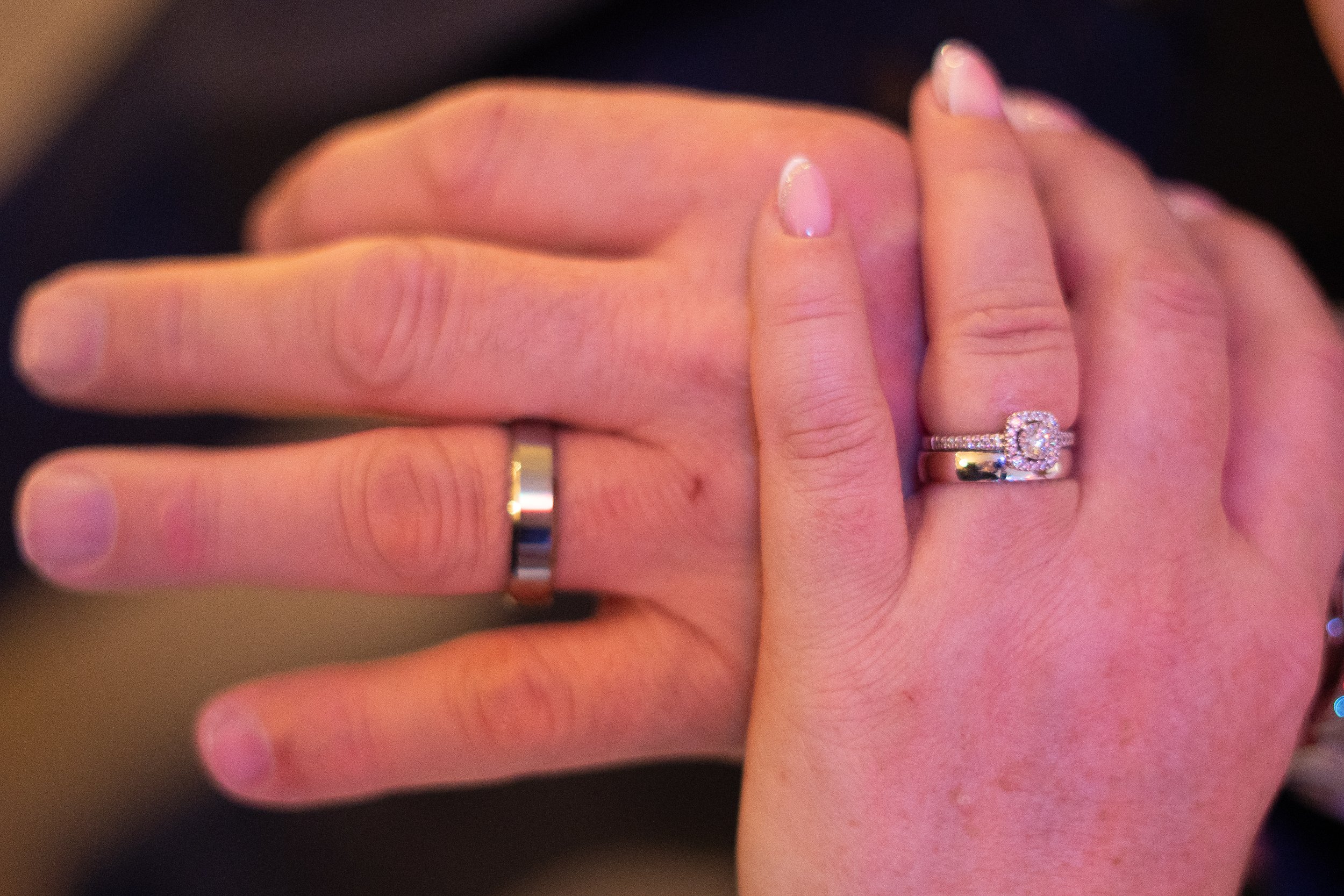 Close-up of two hands showing engagement and wedding rings. The ring finger of the left hand has a shiny silver wedding band, while the right hand has a sparkling engagement ring with a large central stone surrounded by smaller stones, along with a w