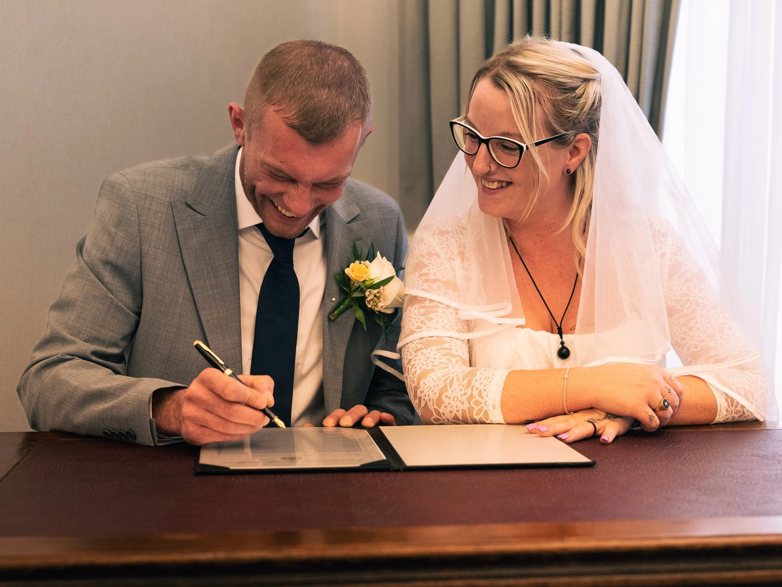 A man in a gray suit and a woman in a white wedding dress signing a document at a table during a wedding ceremony.