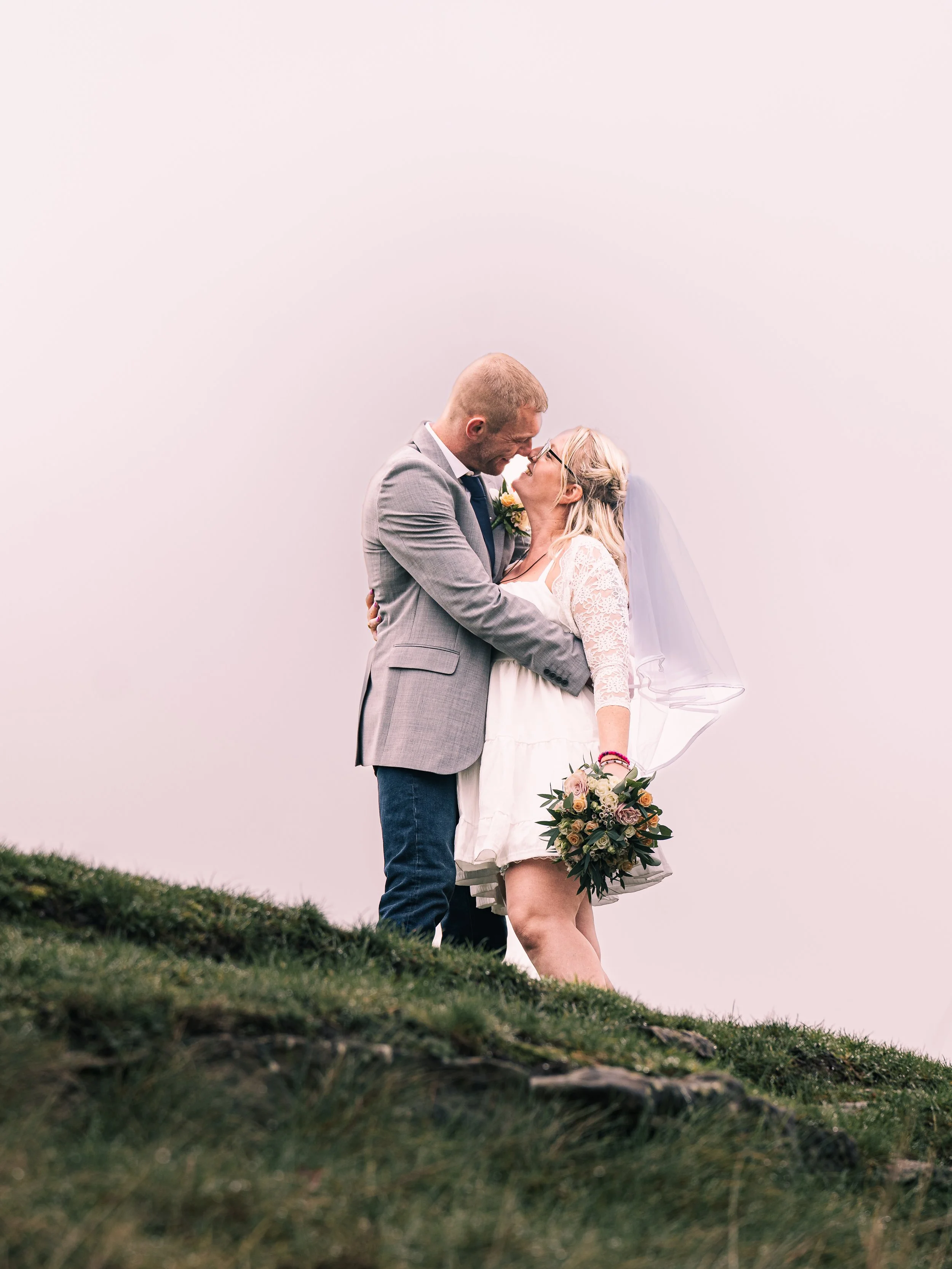 A bride and groom embrace on a grassy hill during their wedding, with the groom wearing a gray suit and the bride in a white dress holding a bouquet, facing each other closely with noses touching.