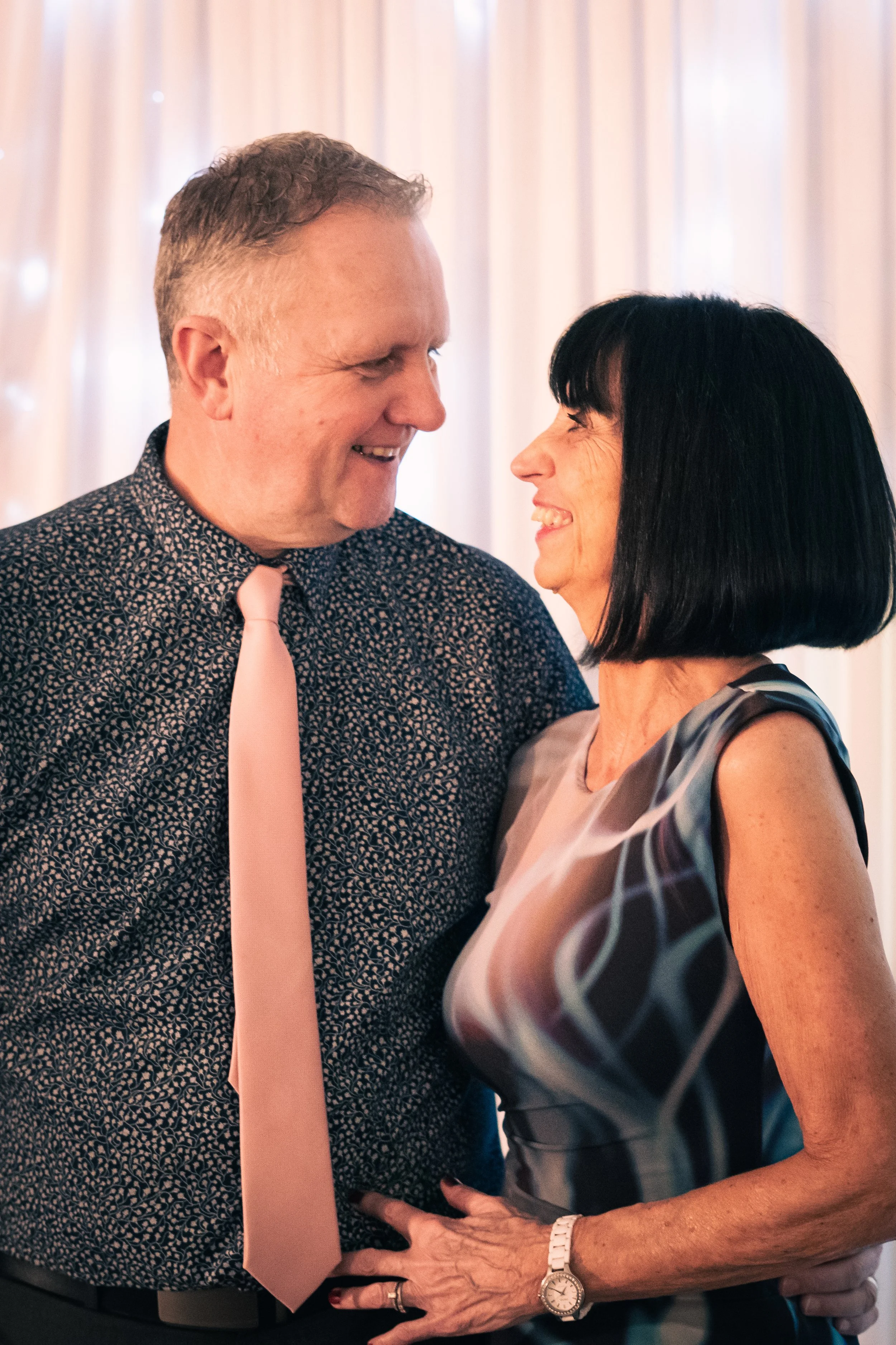 A couple smiling and looking into each other's eyes at a celebration or gathering.
