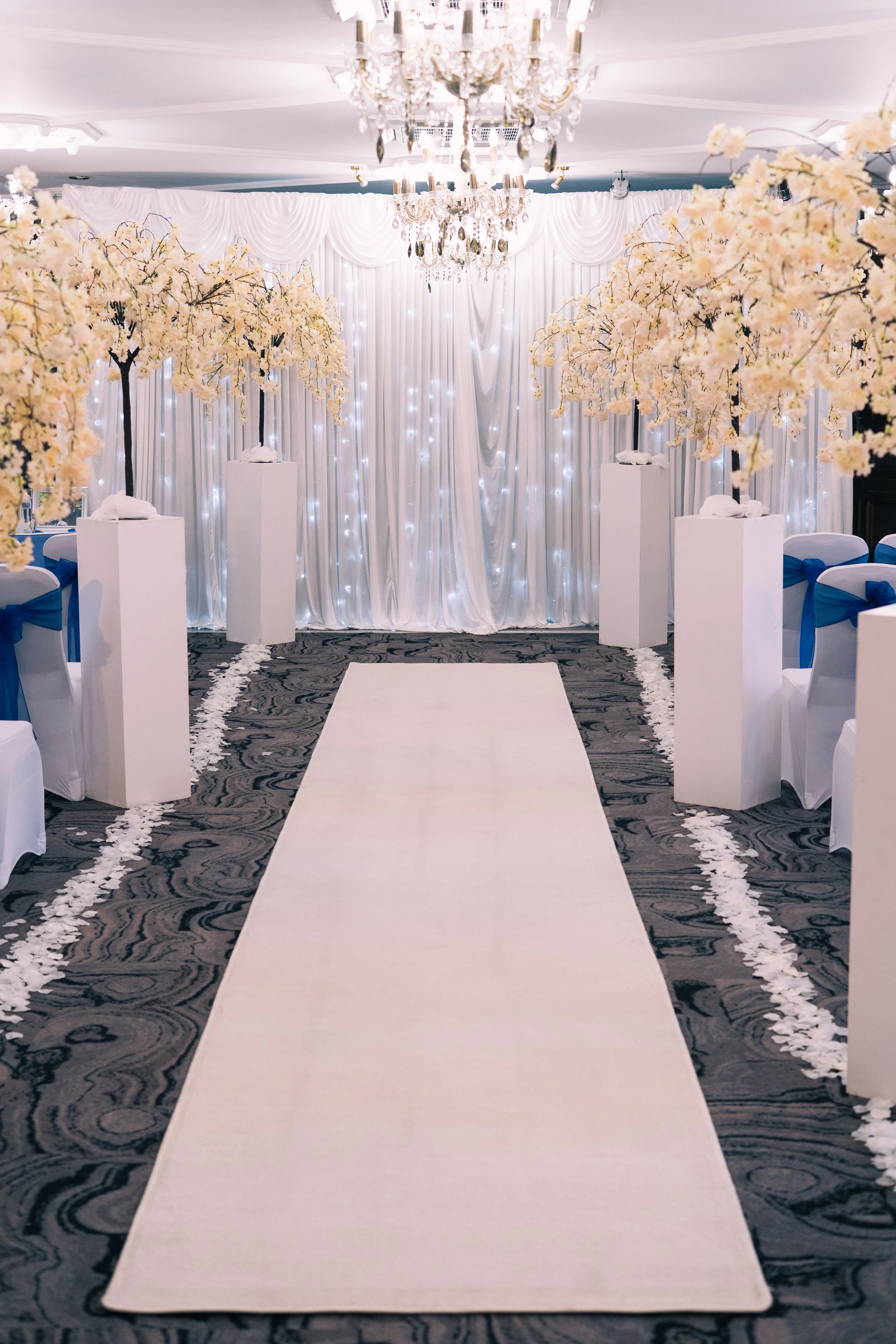 Elegant wedding aisle decorated with white flower petals, lined with tall white pedestals holding white flowering trees, leading to a backdrop with white drapes and twinkling lights, illuminated by a chandelier.
