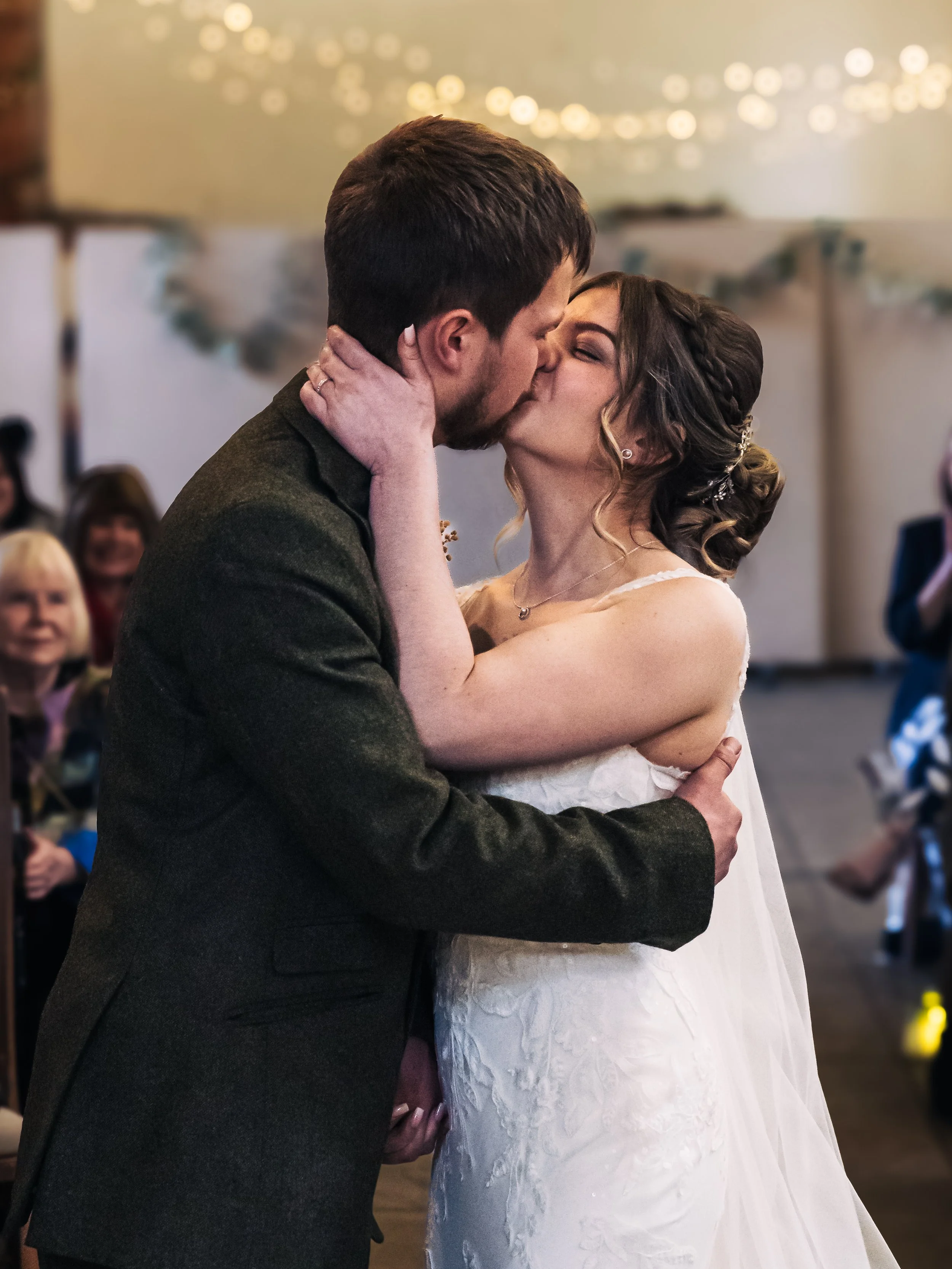 A bride and groom share a kiss during their wedding ceremony, with guests watching in the background.