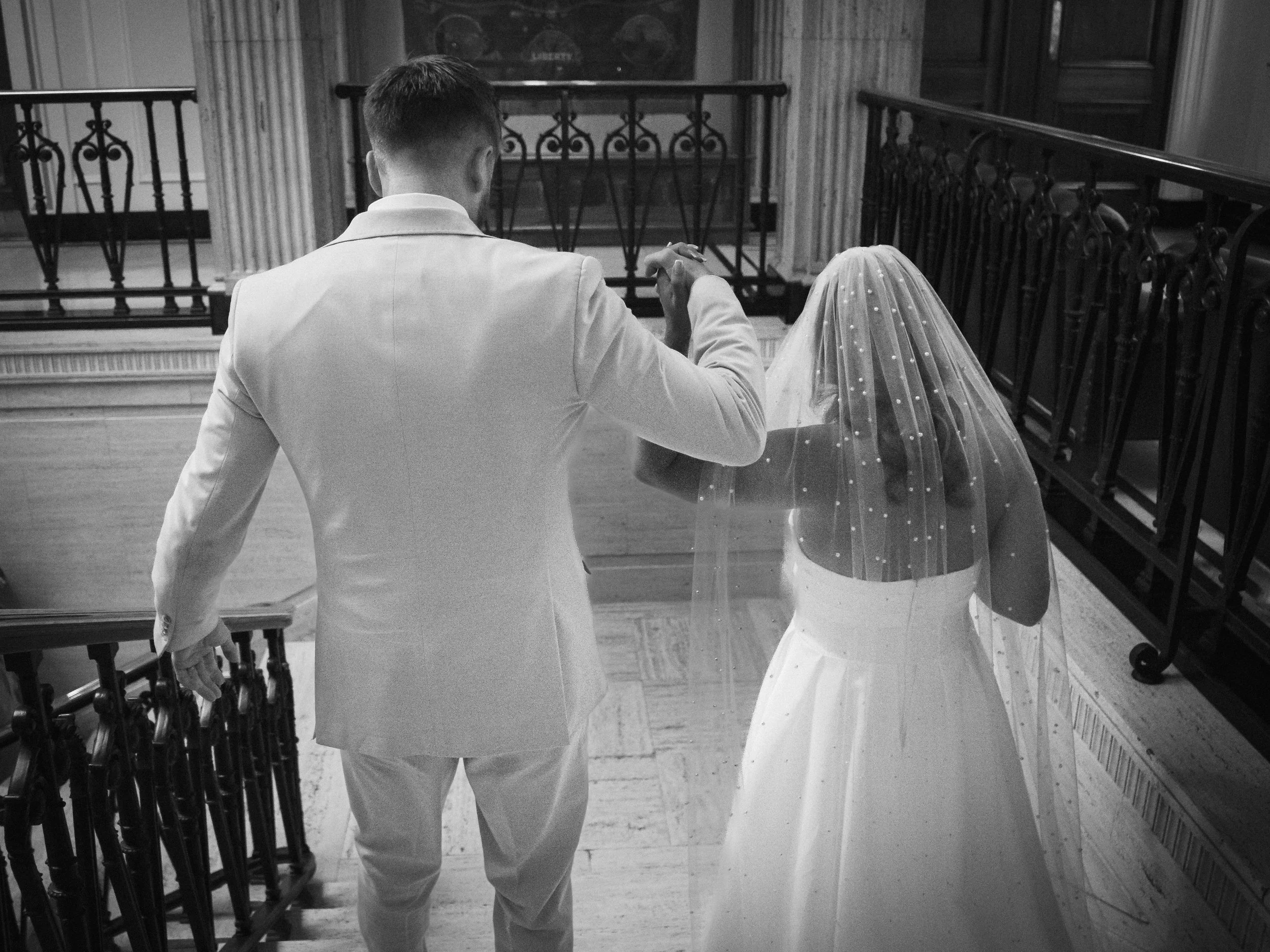 A black-and-white photo of a bride and groom at a wedding ceremony, with the groom holding the bride's hand as she wears a wedding dress and veil, on a staircase inside a church or historic building.