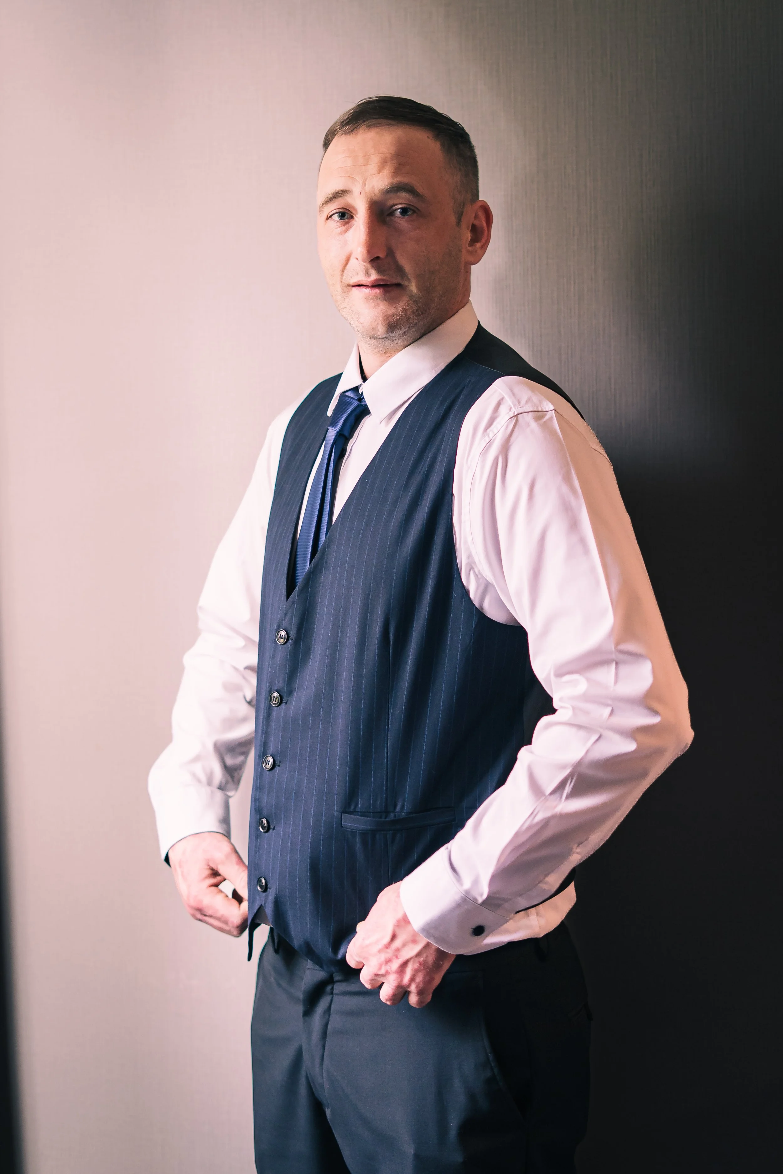 A man in business attire, wearing a white shirt, dark blue vest, and striped tie, standing against a wall with a neutral expression.