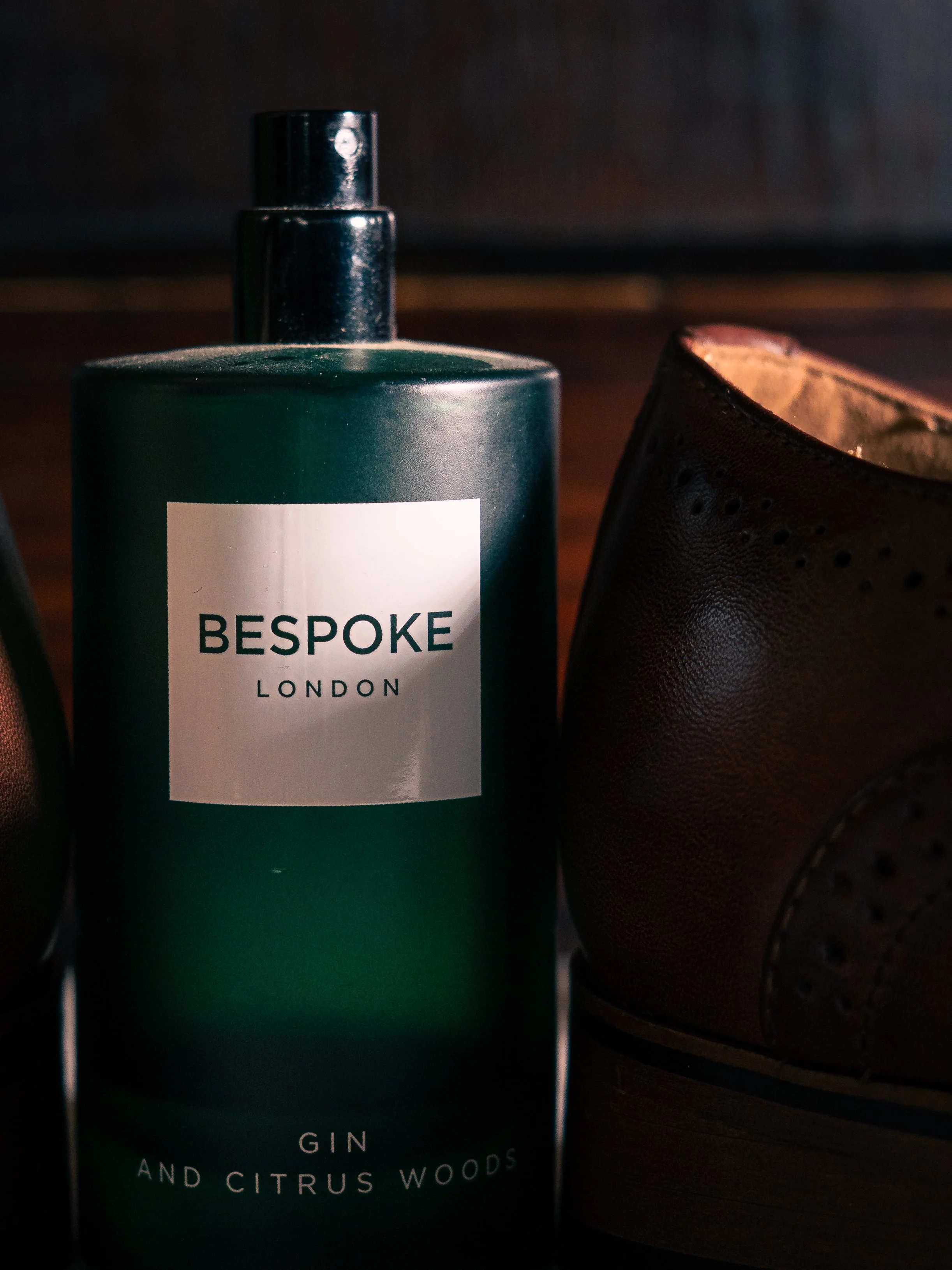 A bottle of Bespoke London gin with a label that reads 'GIN AND CITRUS WOODS,' positioned next to a brown leather shoe on what appears to be a wooden surface.