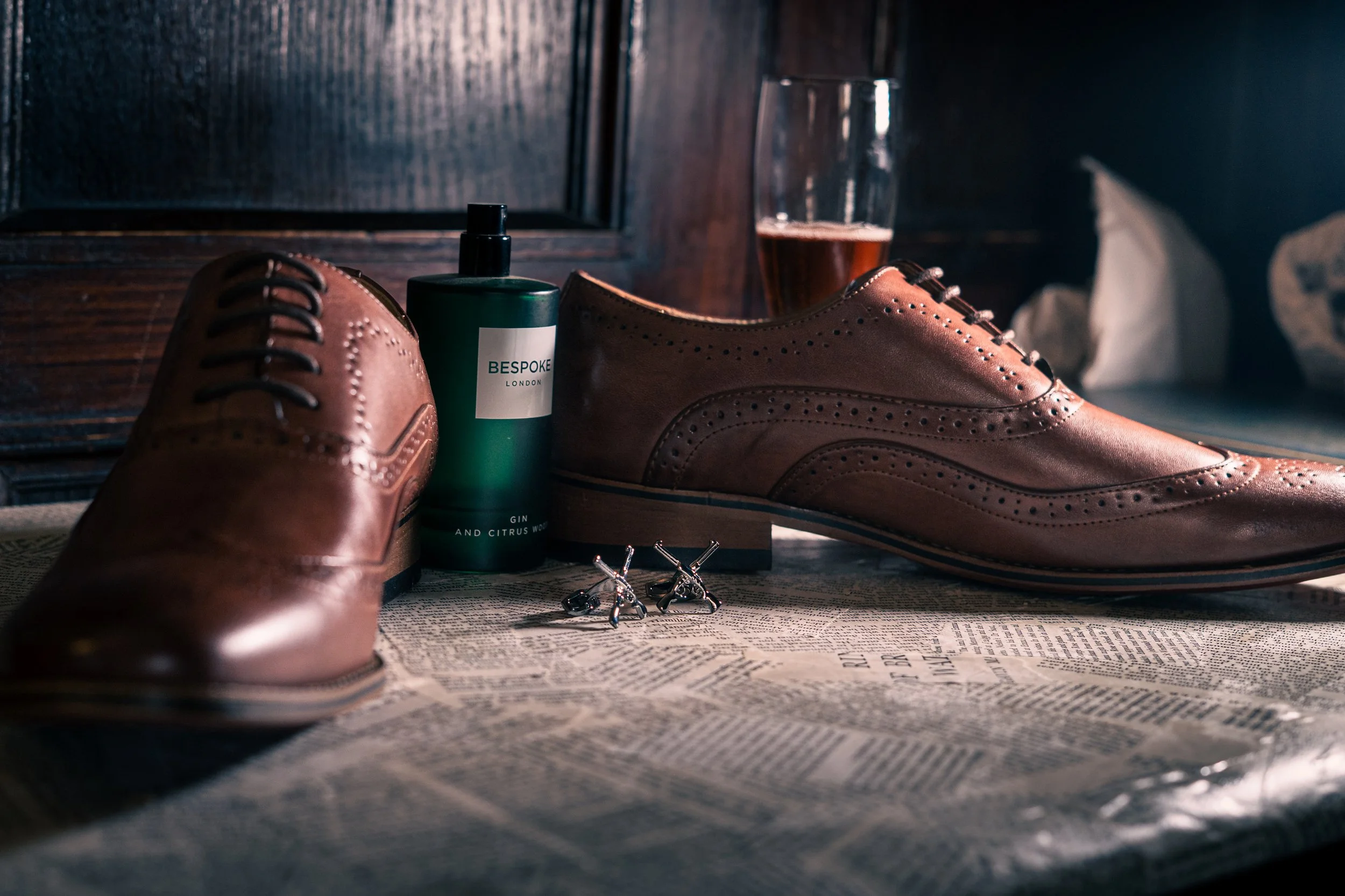 Brown leather dress shoes, a bottle of shoe polish, a glass of beer, a newspaper, and a pair of cufflinks on a table with a dark wood background.
