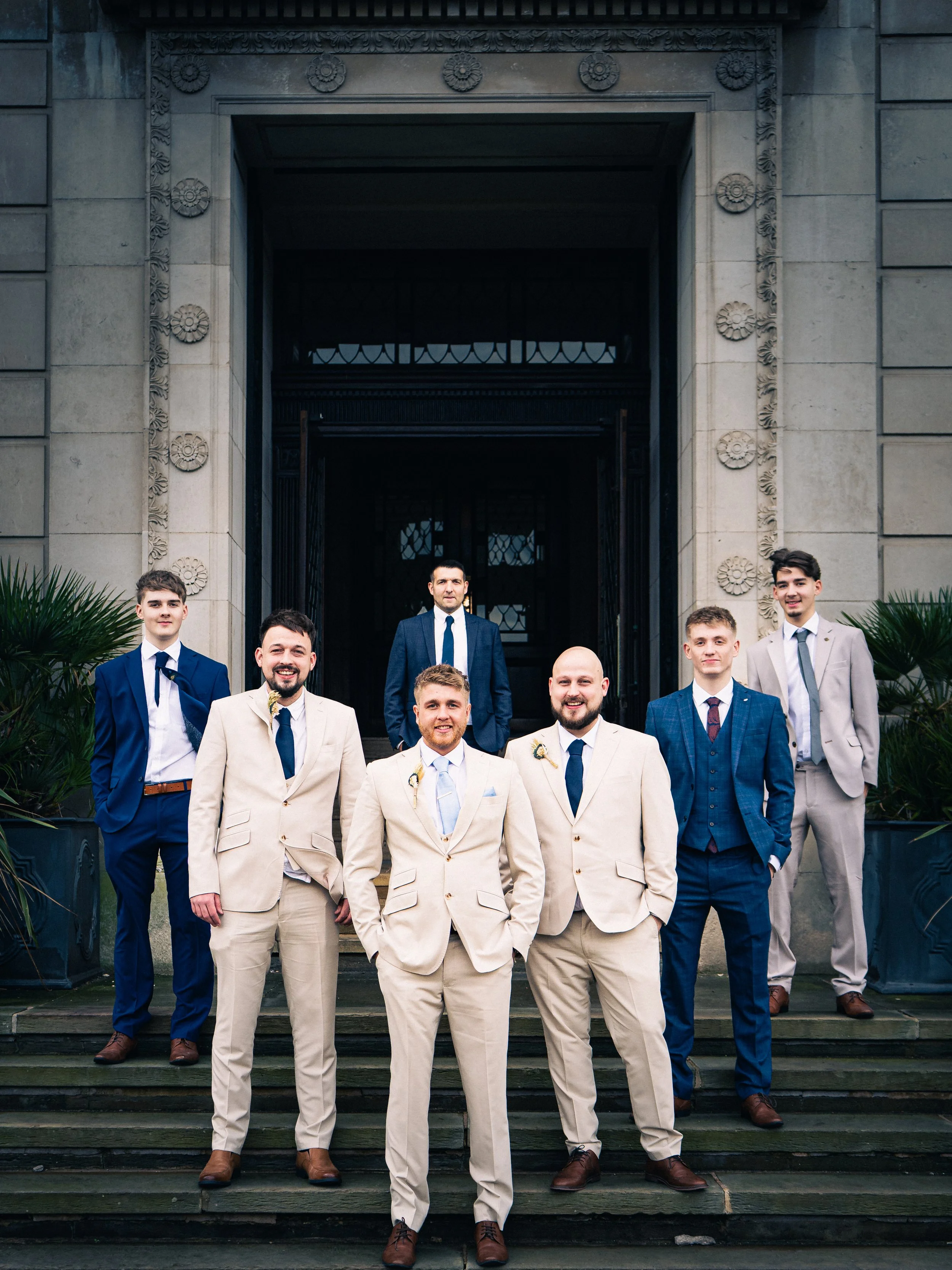 Group of seven men in formal suits standing on steps outside a building with stone walls and decorative carvings, posing for a photo.