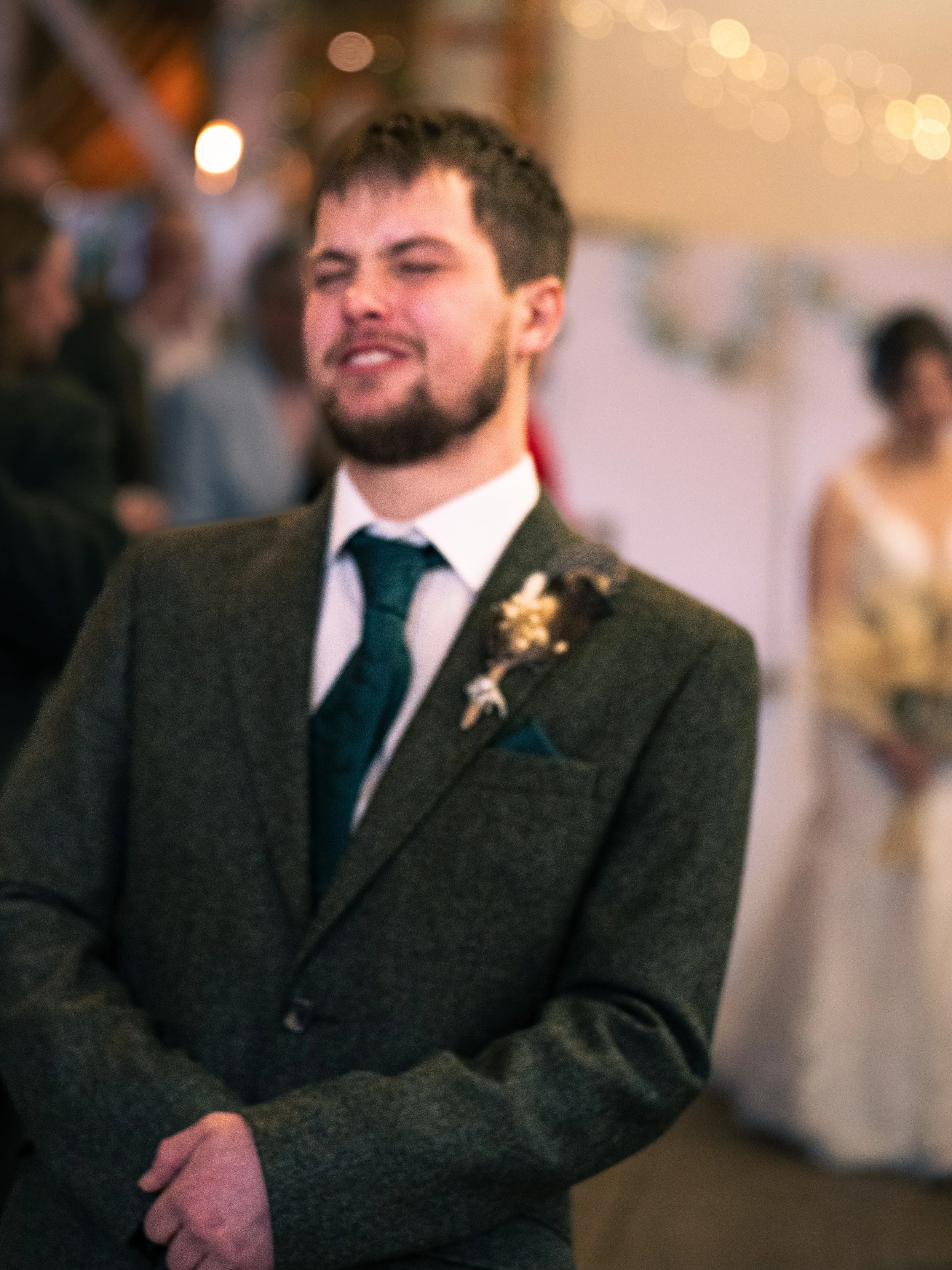 A man in a suit with a boutonniere standing at a wedding reception, smiling with his eyes closed as others dance and socialize in the background.