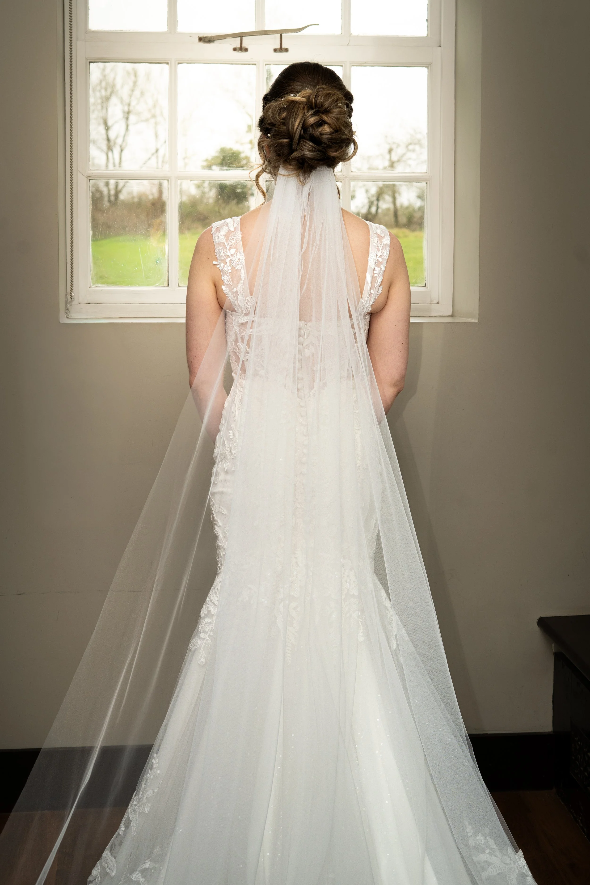 A bride in a white wedding dress with lace details and a long veil is standing indoors, facing a window with a view of green grass and trees outside.