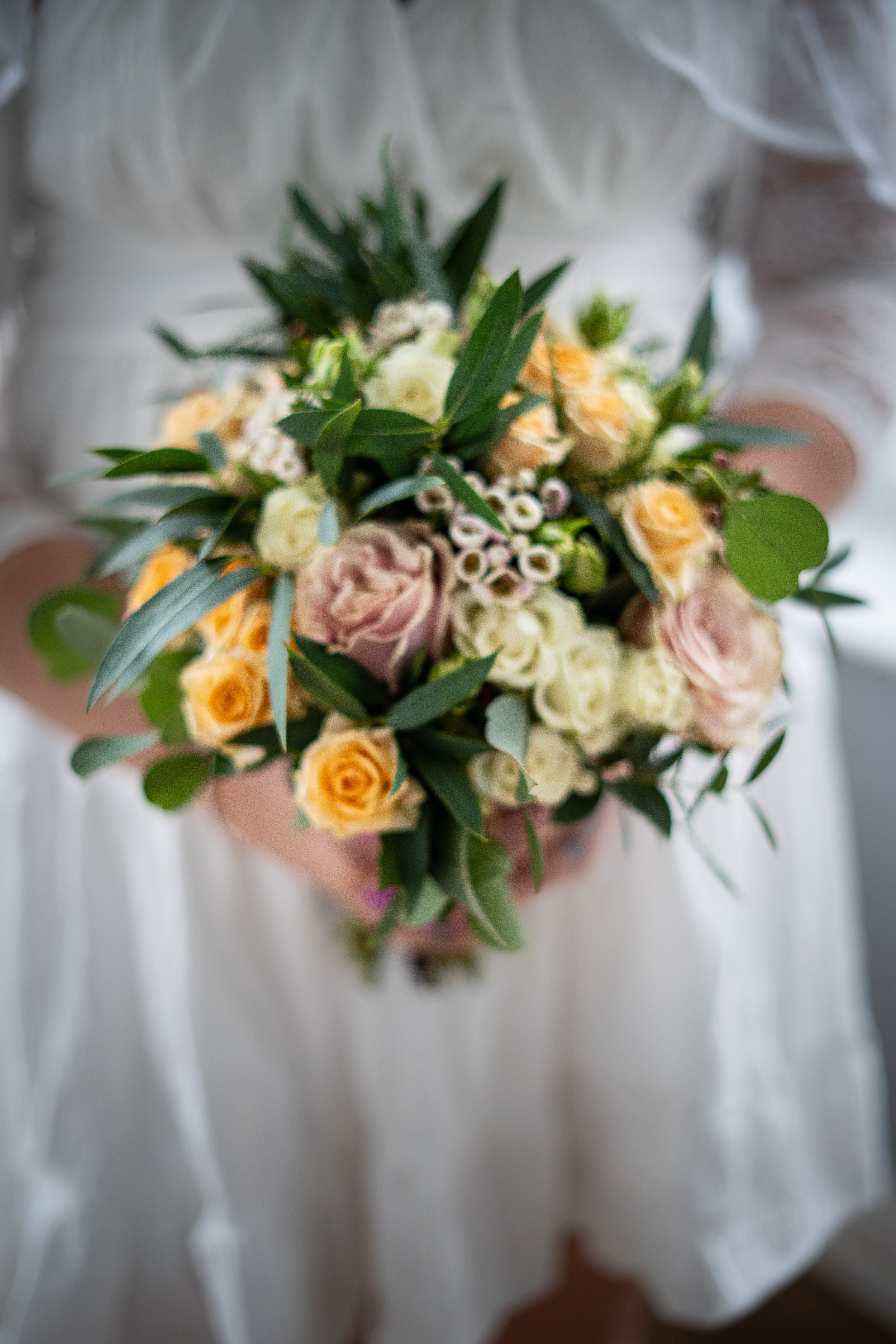 A person in a white dress holding a bouquet of light pink, white, and yellow flowers with green leaves.