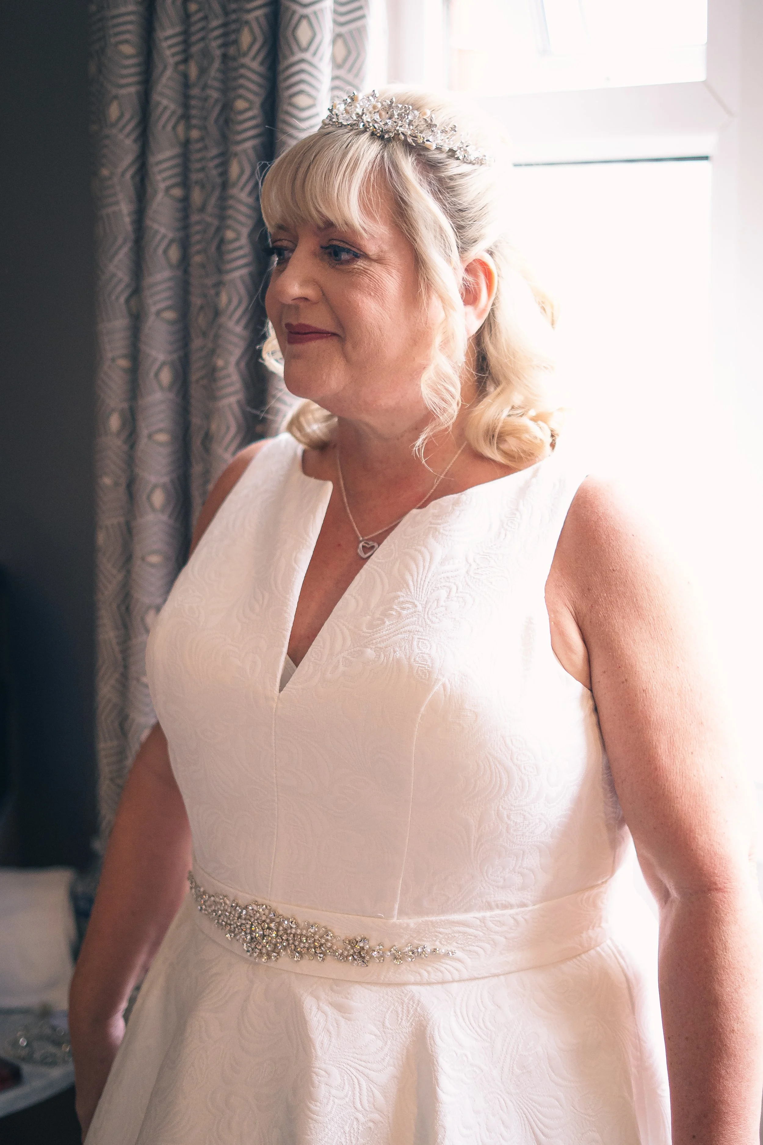 A woman wearing a white dress and a tiara, standing near a window, preparing for a special occasion.