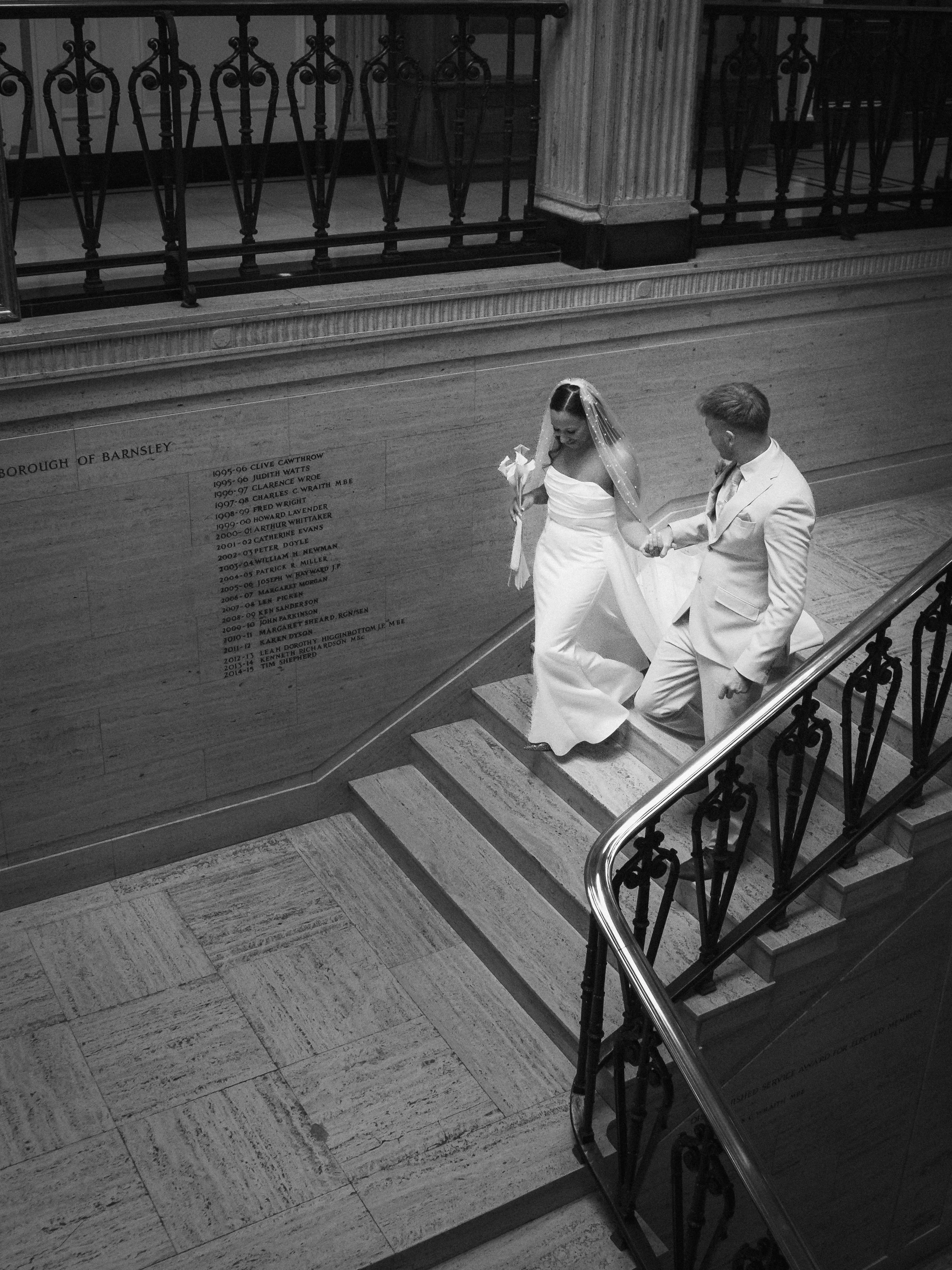 Black and white photo of a bride and groom ascending a staircase inside a building, holding hands. The bride is in a wedding dress and veil, holding a bouquet. The groom is in a suit.