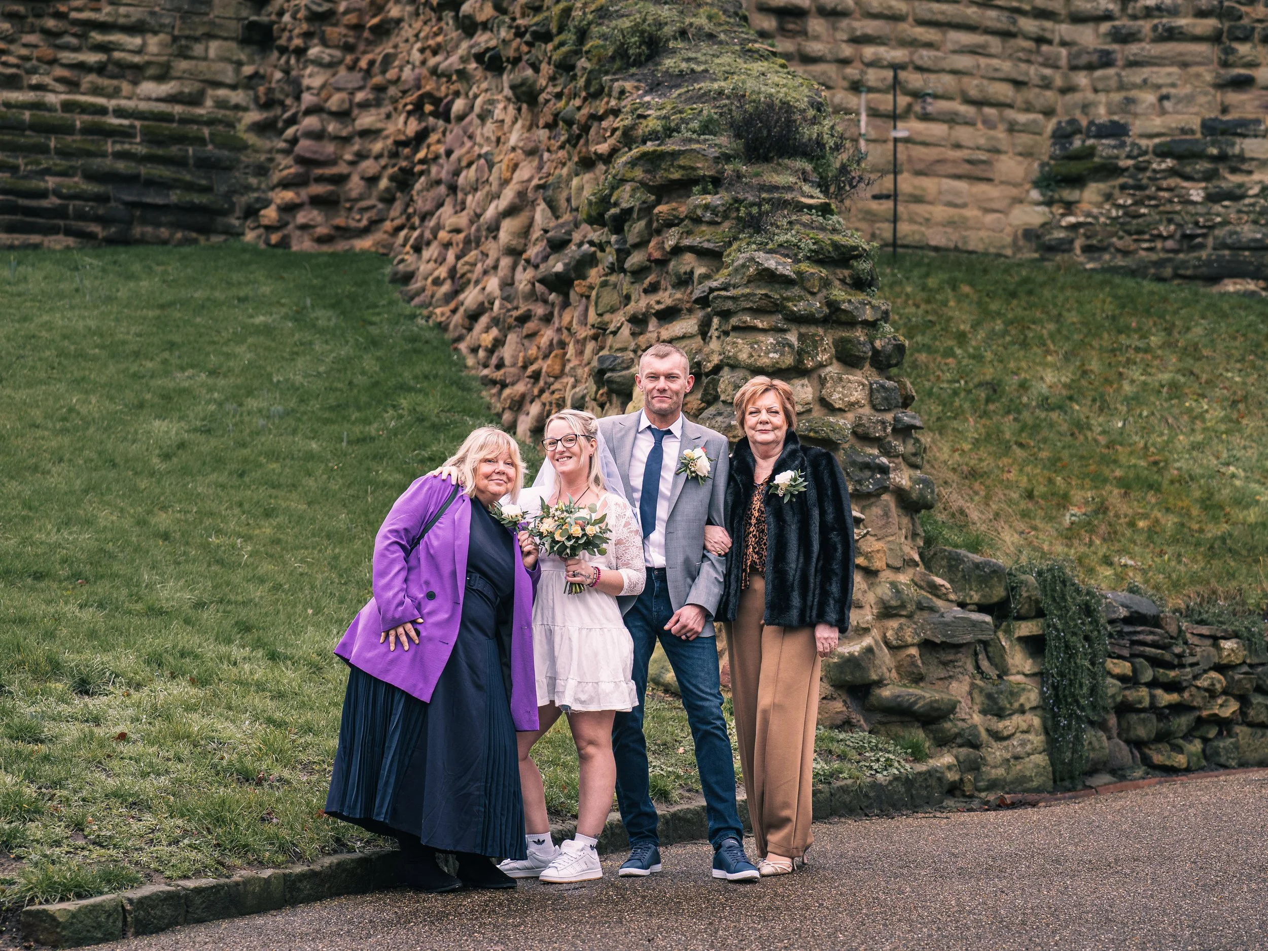 A group of five people, including a bride holding a bouquet, standing outdoors in front of a stone wall. They are dressed semi-formally, suggesting a wedding celebration.