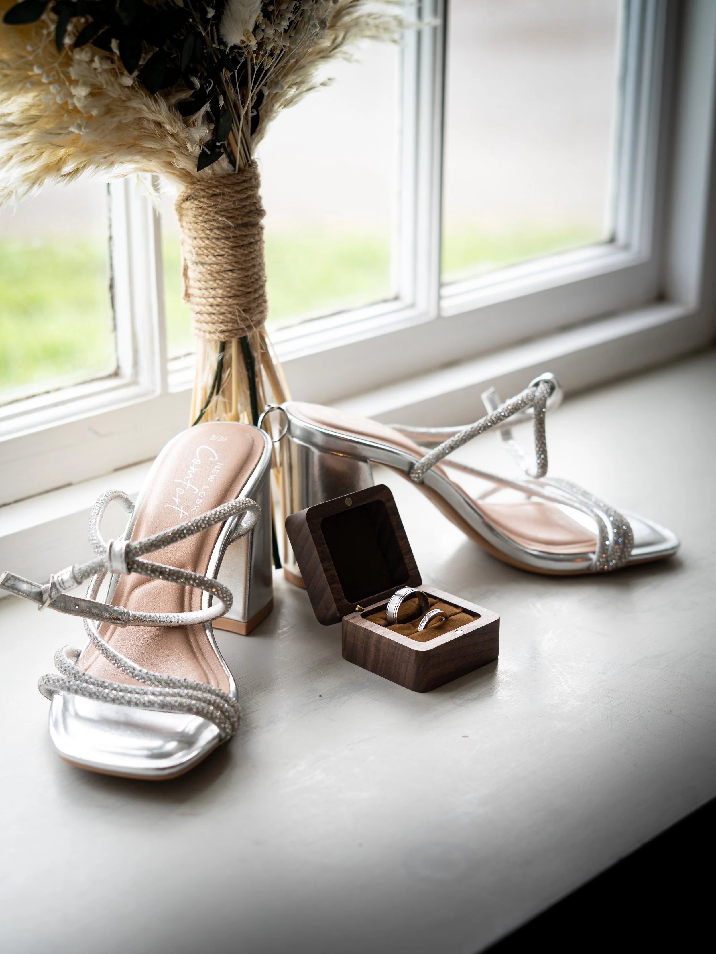 A pair of silver high-heeled sandals with rhinestone embellishments, a small wooden box containing wedding rings, a bouquet of dried flowers, and a window background.