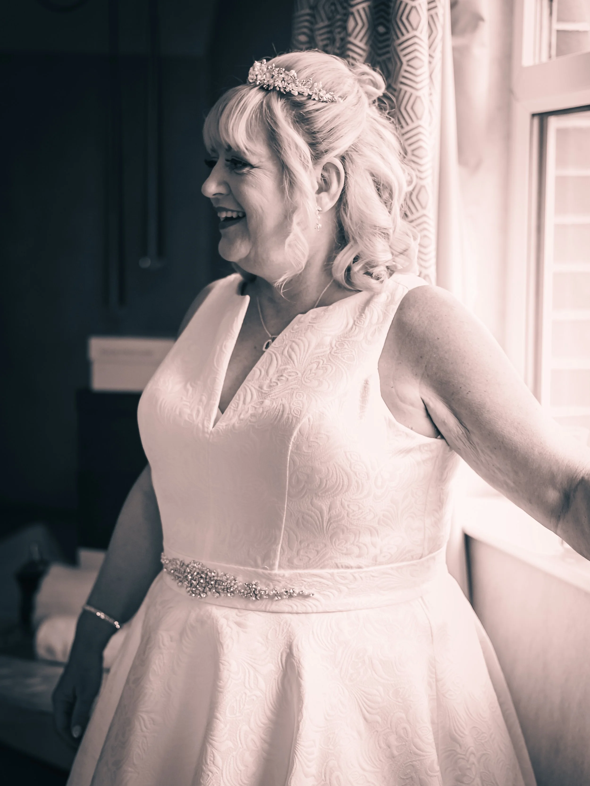 A woman in a wedding dress, standing near a window, smiling and gazing to her left, with a tiara and jewelry, in a softly lit room.