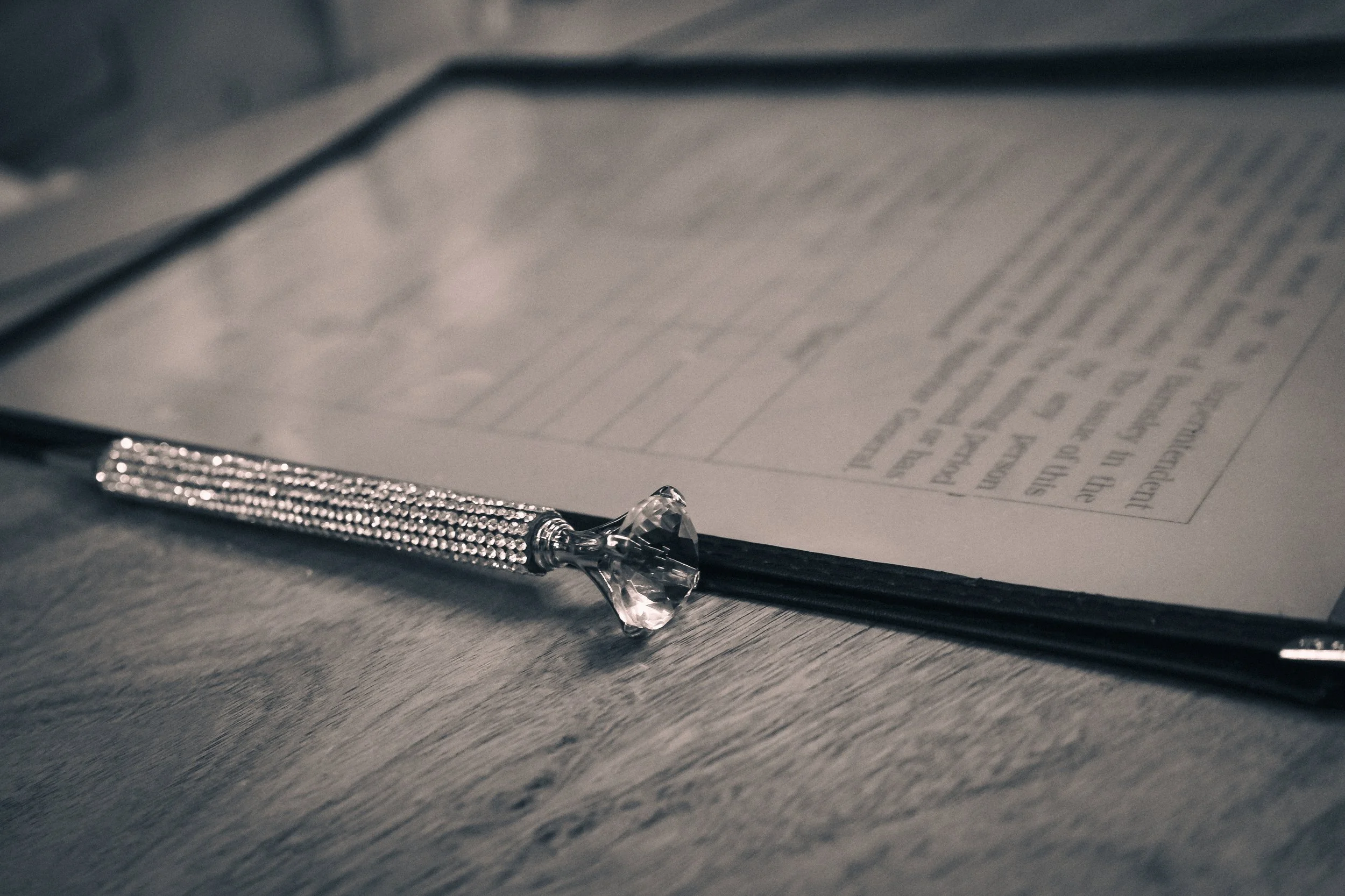 A pen with a silver, bead-like textured handle and a large, faceted clear gemstone at the top, resting on an open book or document on a wooden surface.