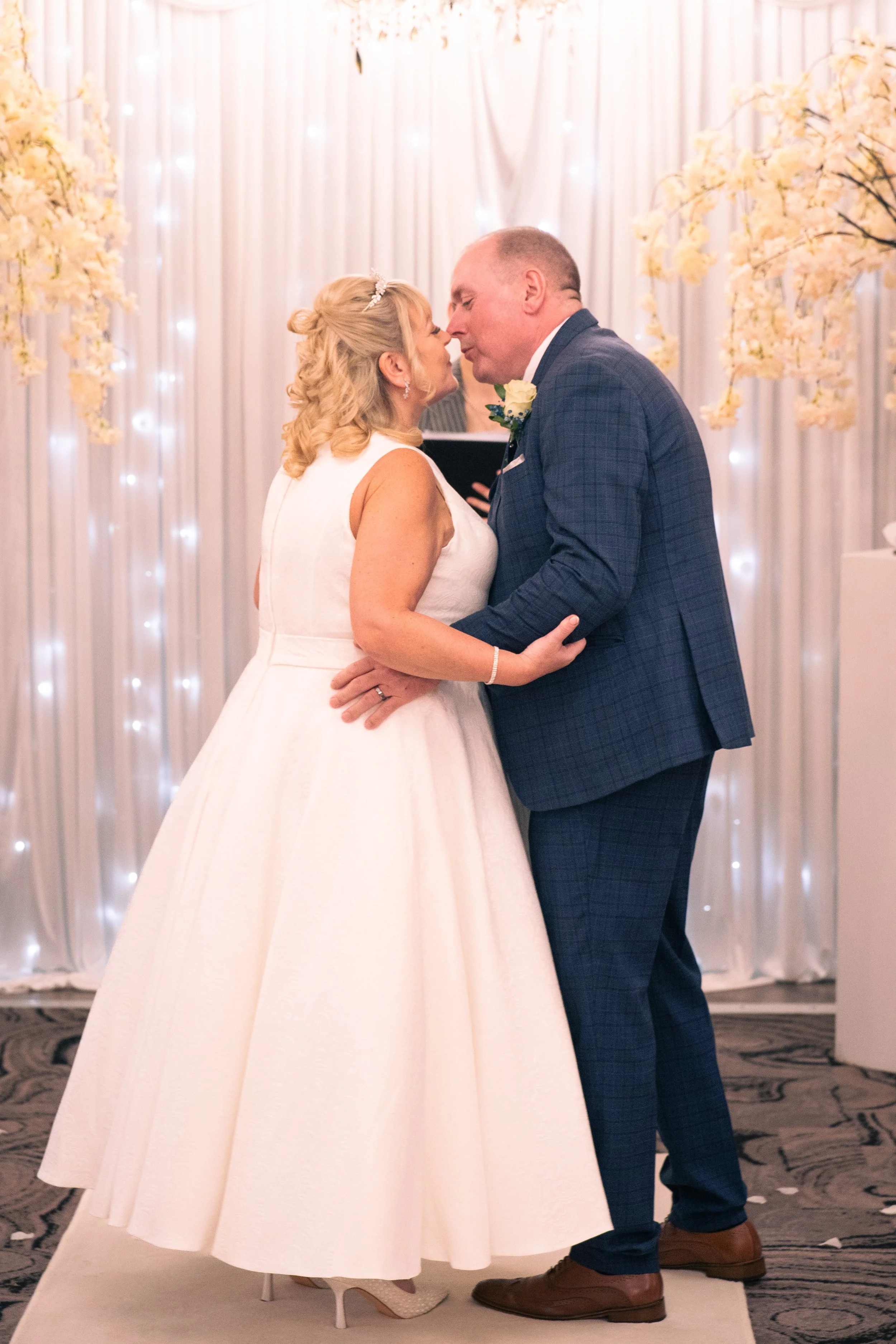 A bride and groom share a kiss during their wedding ceremony, with the bride in a white dress and the groom in a blue suit, in front of a decorated backdrop with flowers and lights.