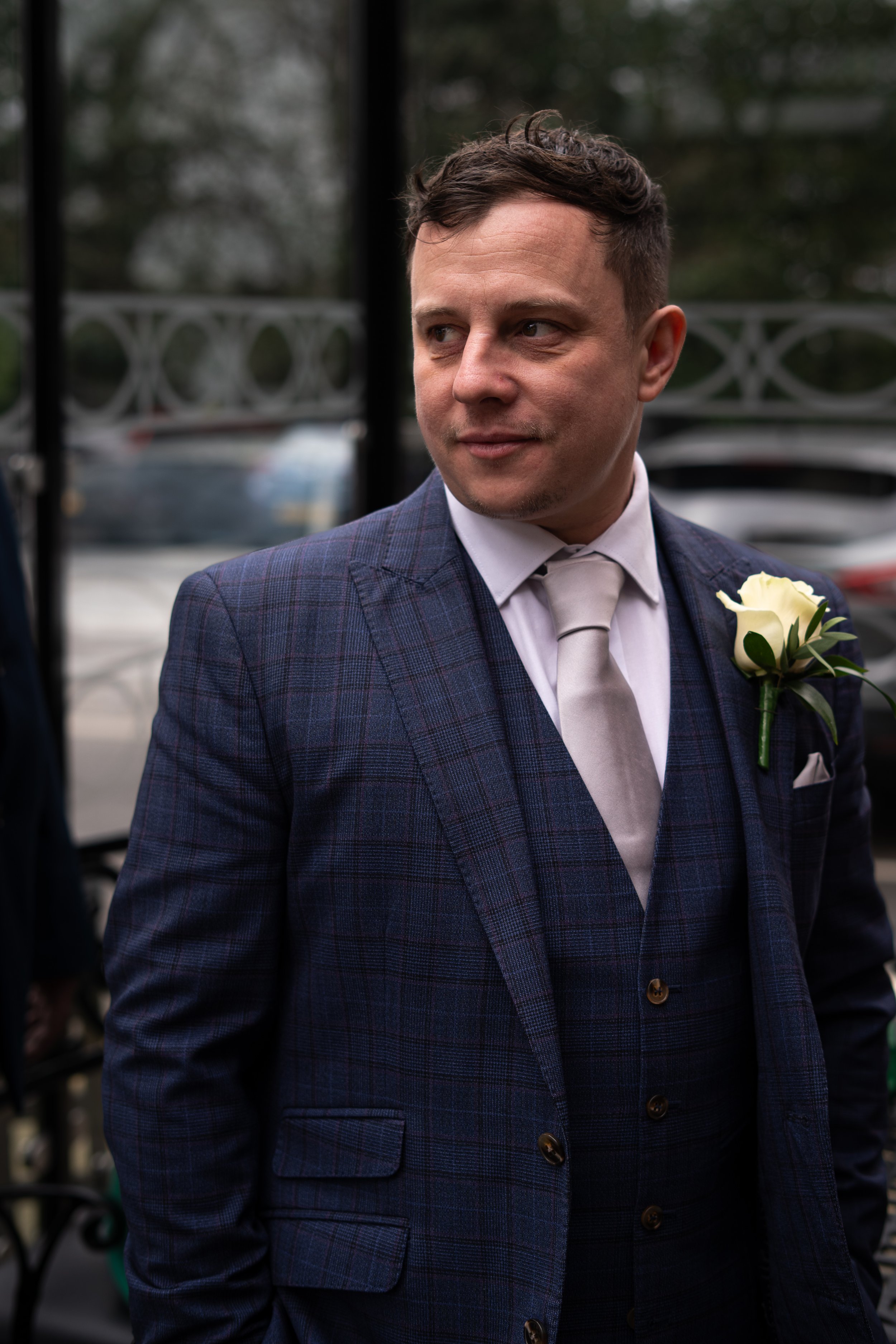 A man dressed in a plaid blue suit with a white shirt and a light-colored tie, wearing a white rose boutonniere, stands outdoors near cars.