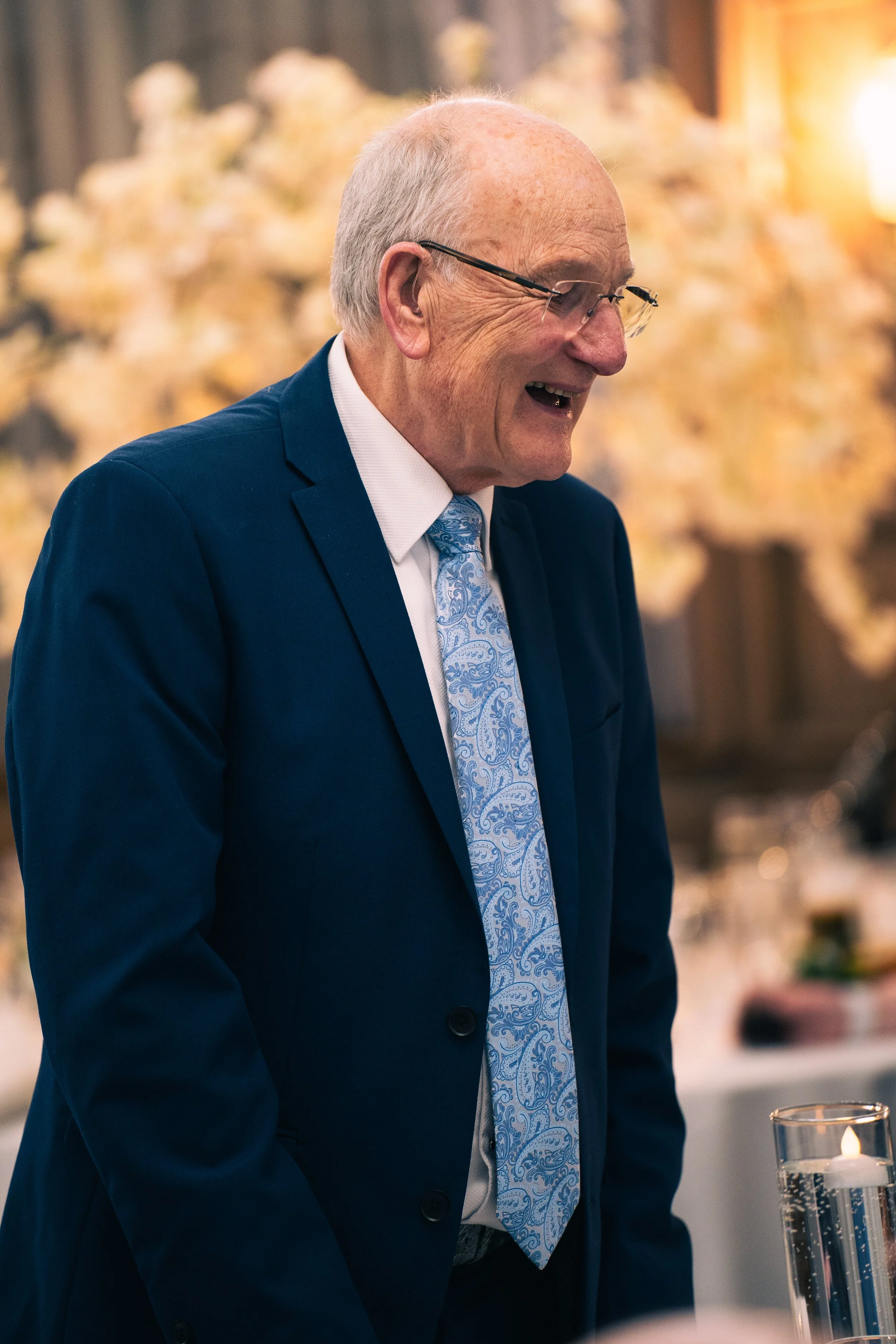 Older man with glasses, wearing a navy blue suit and a light blue paisley tie, smiling and looking to his right at an indoor event with warm lighting and blurred floral decorations in the background.