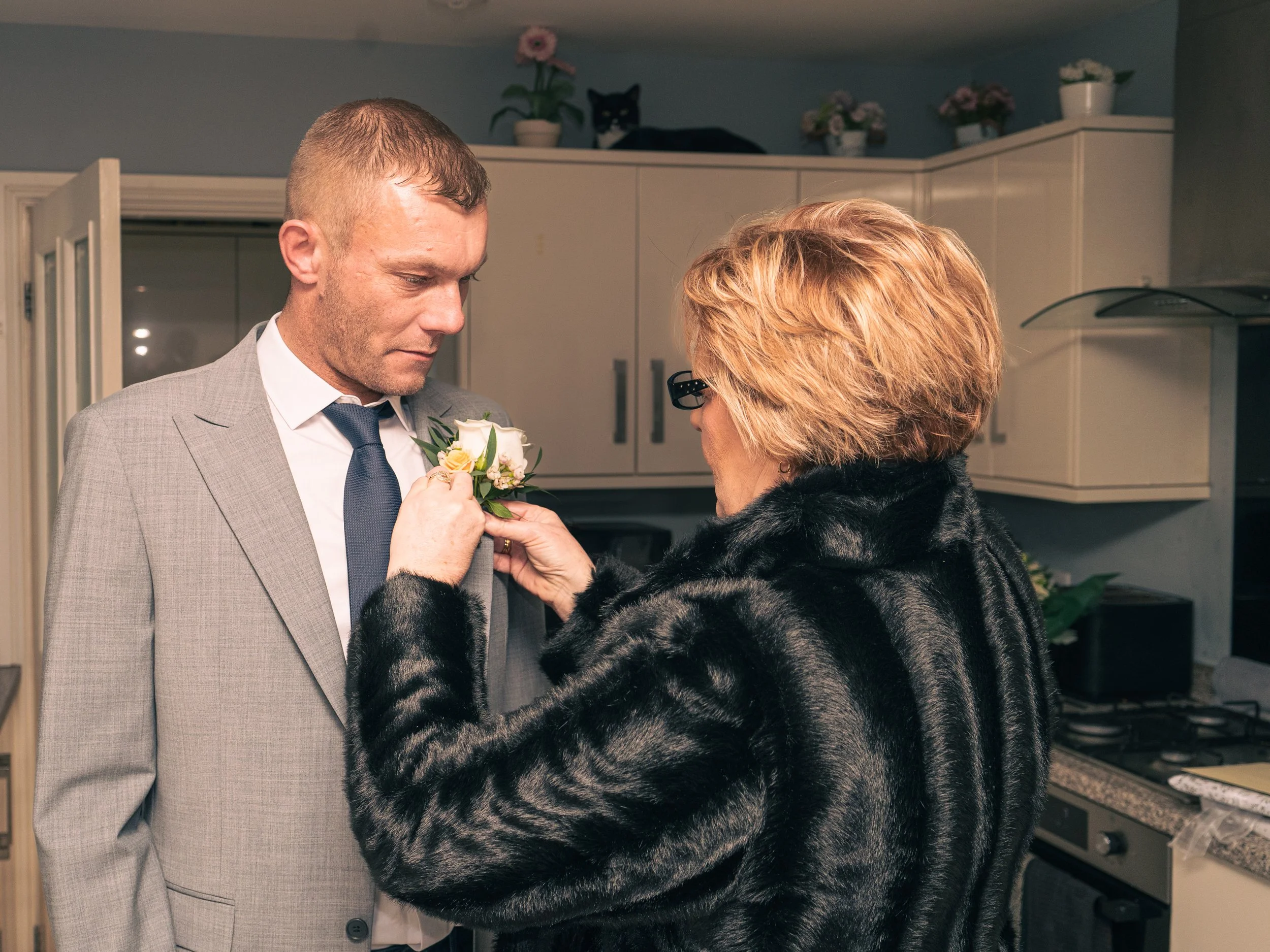 An older woman pinning a boutonniere on a young man in a kitchen.
