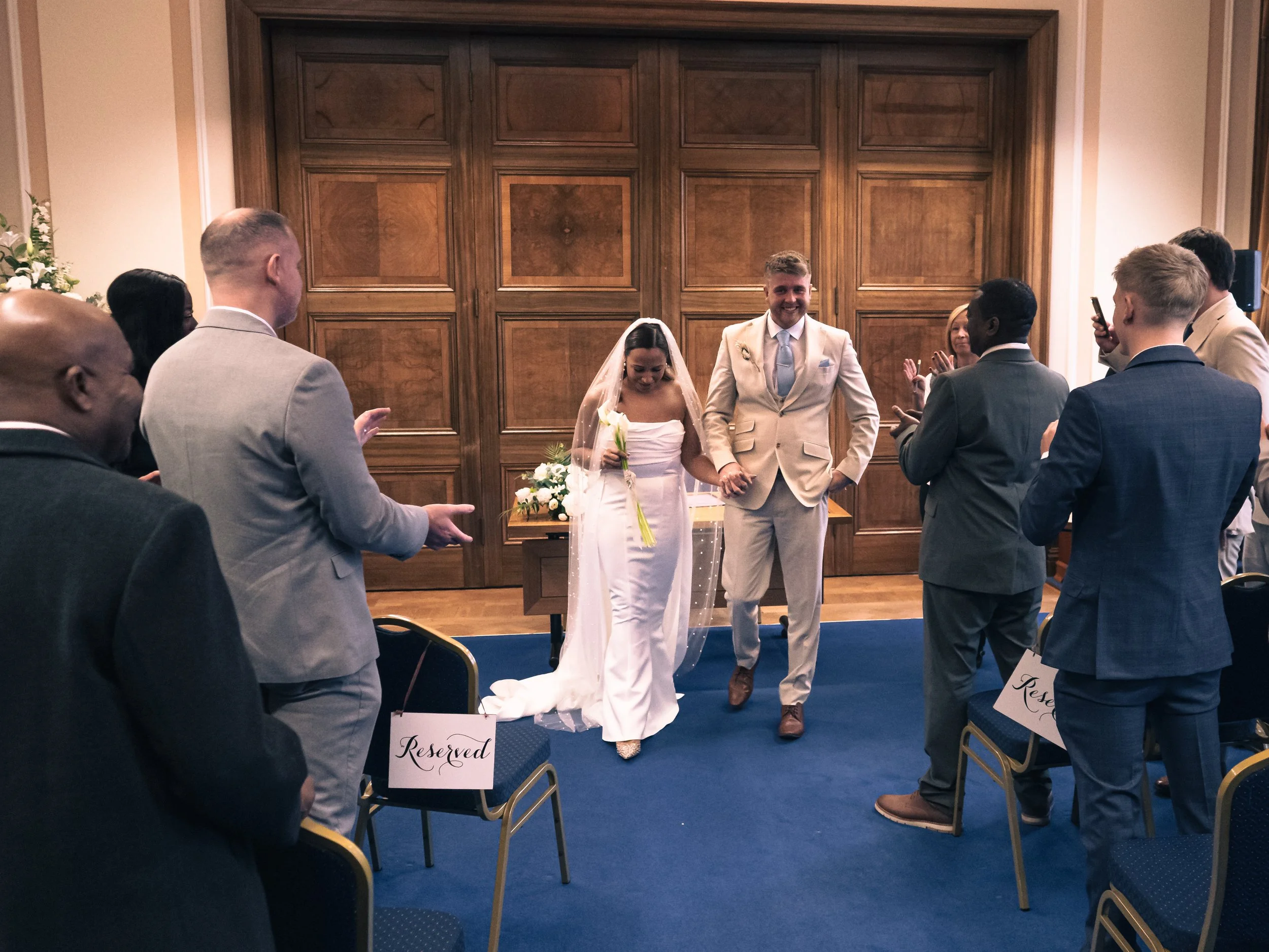 A bride and groom walking down the aisle holding hands at their wedding ceremony. The bride is holding a bouquet, and the groom is dressed in a light suit. Guests are standing and clapping, some taking photos.