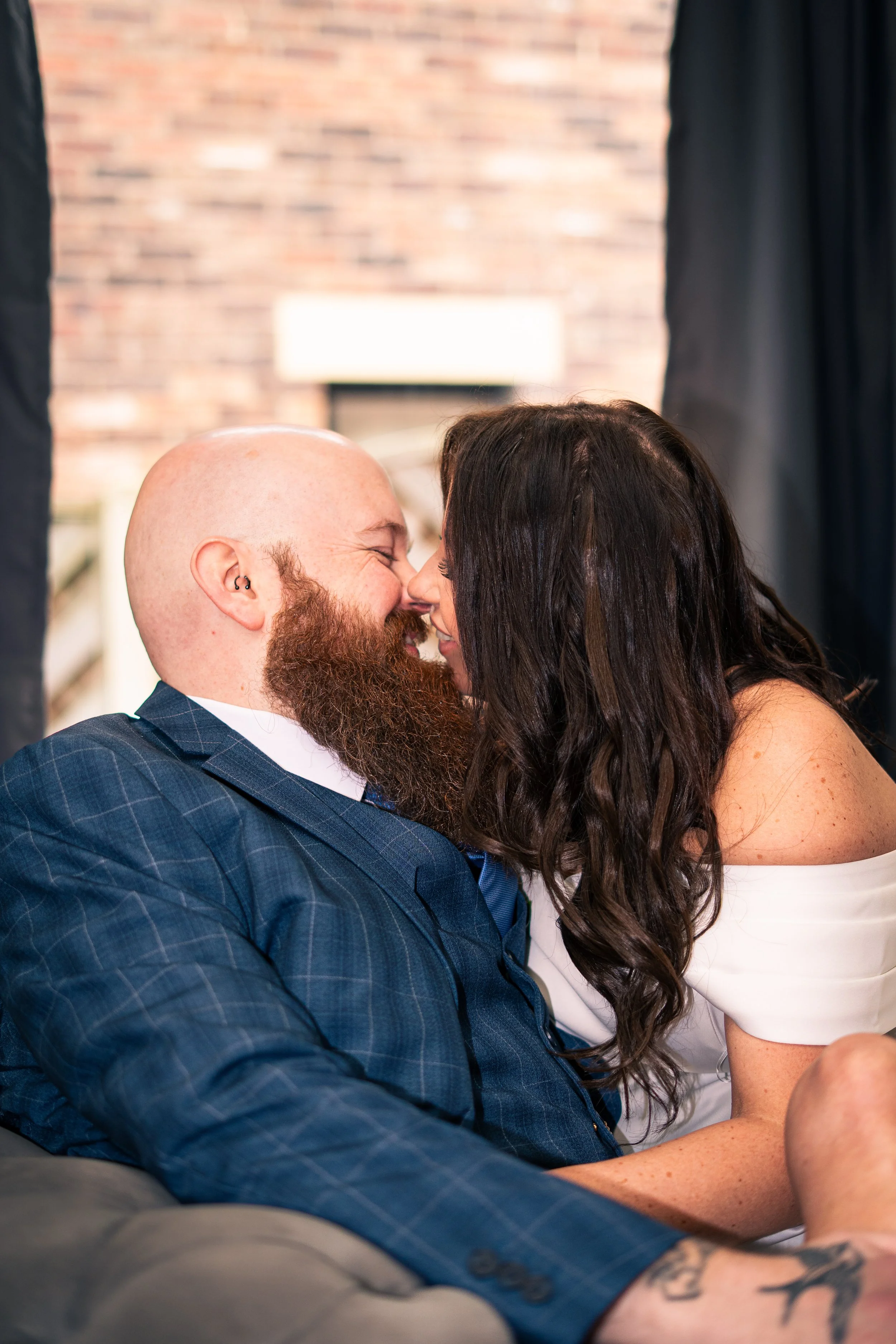 A couple with their noses touching, smiling and about to kiss, indoors with curtains and a brick wall in the background.