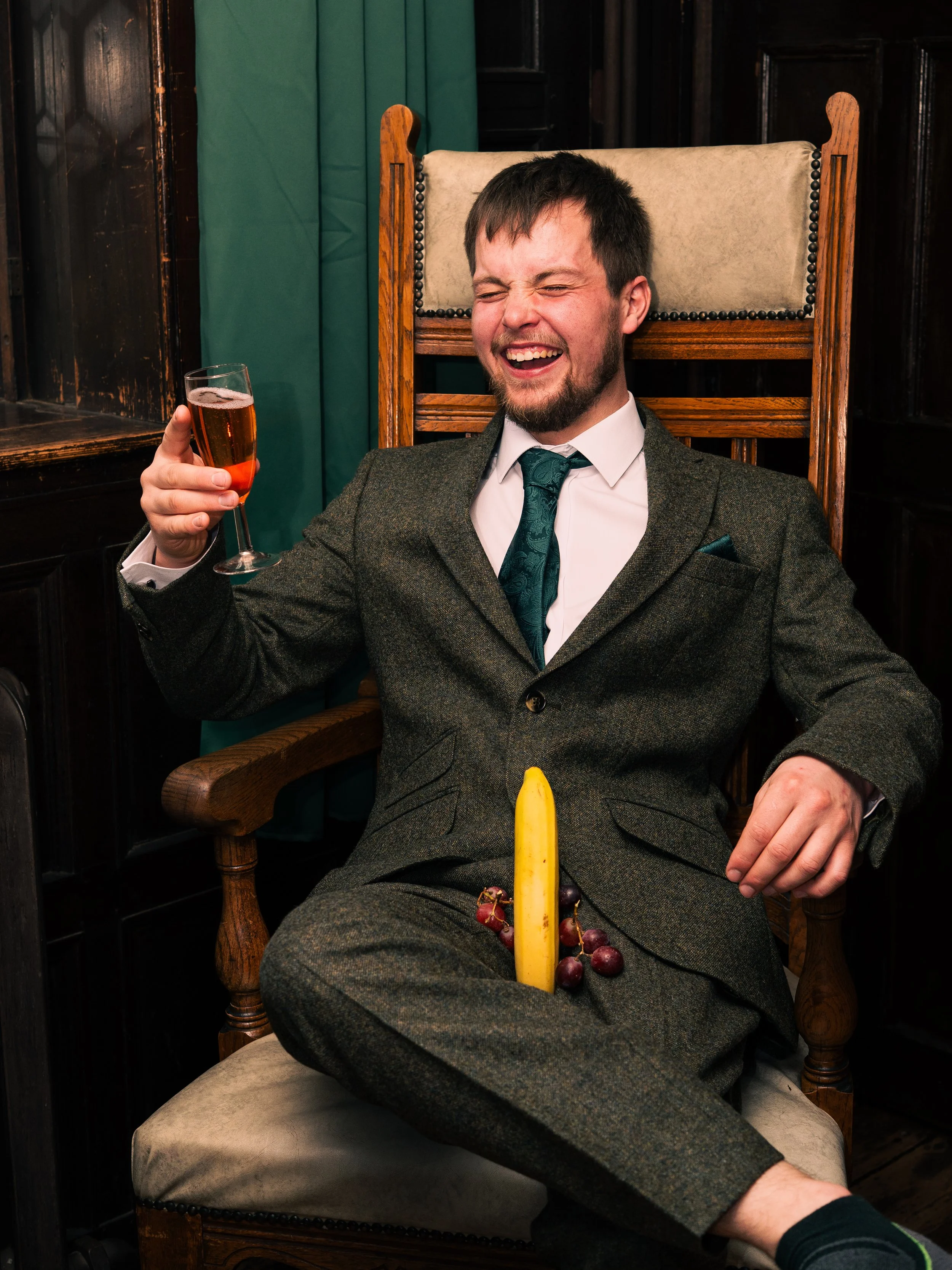 Man sitting in a wooden chair, laughing, with a banana, grapes, and a vintage-style suit, in a dimly lit room with dark wood paneling, holding a glass of wine.