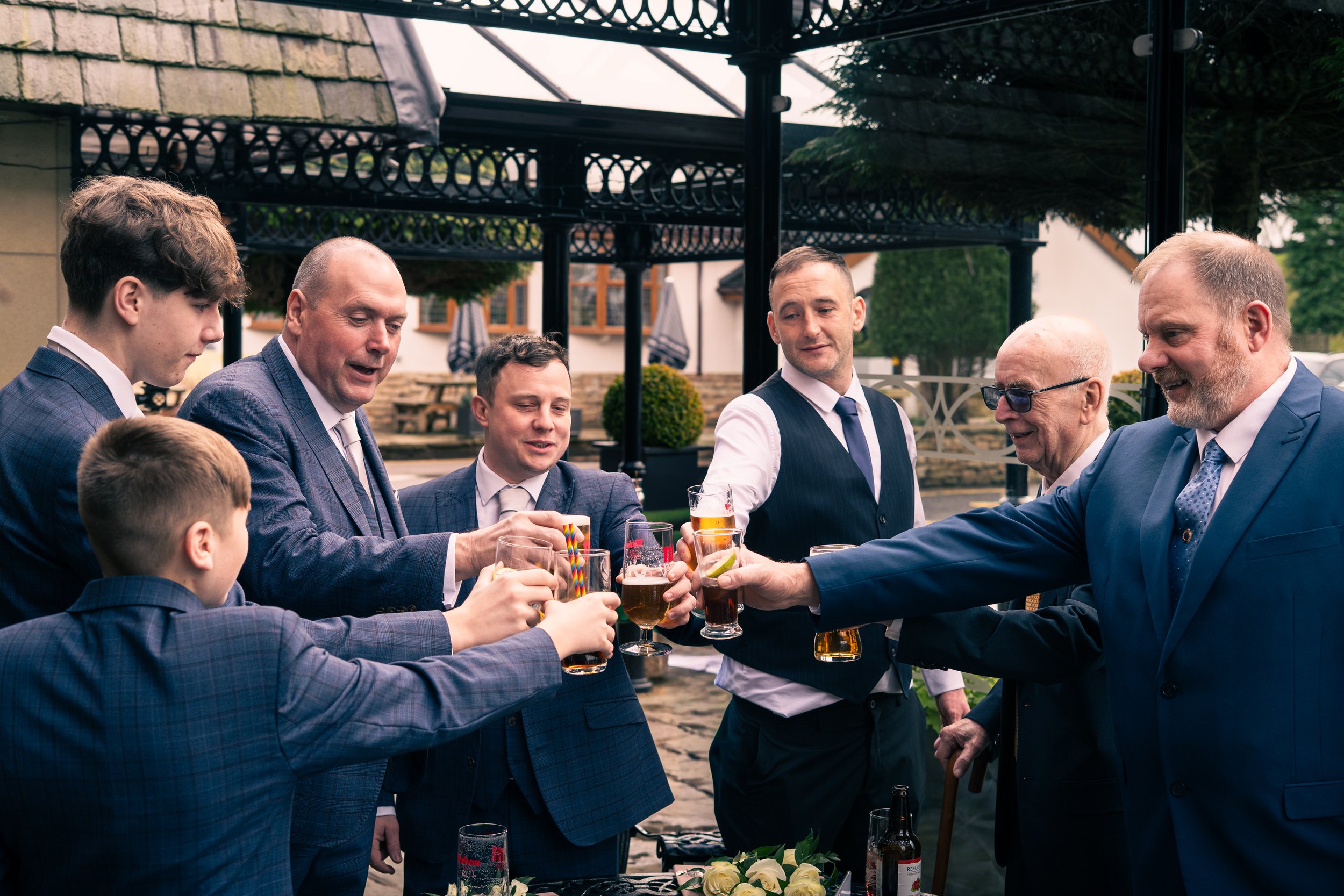 Group of men and boys in suits gathered outdoors raising glasses in a toast.