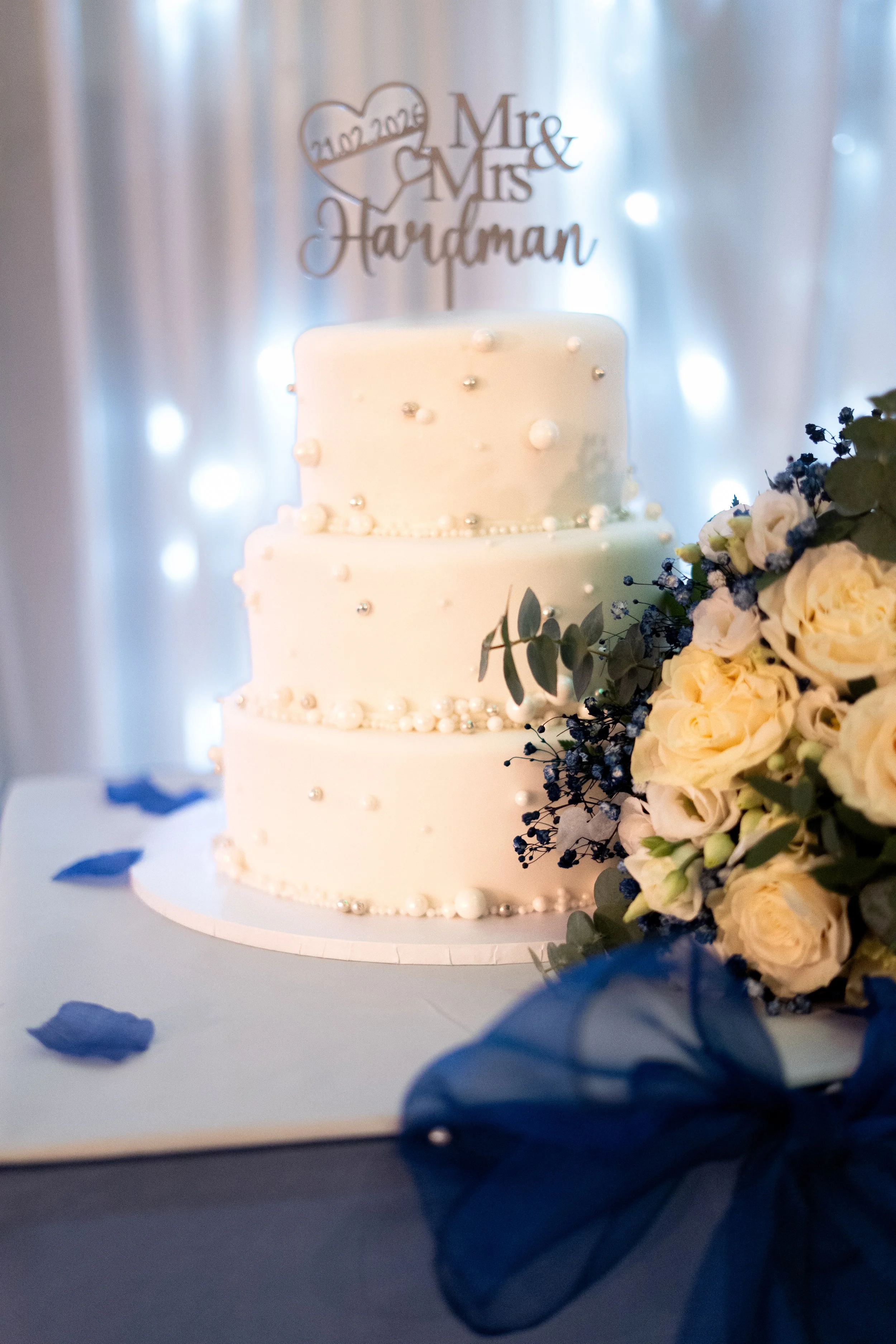 A three-tiered white wedding cake decorated with small pearl-like and silver balls, with a wedding topper that says "Mr. & Mrs. Handman" and a heart with the date "21.02.2023" above it, surrounded by a bouquet of white roses and greenery, set on a ta