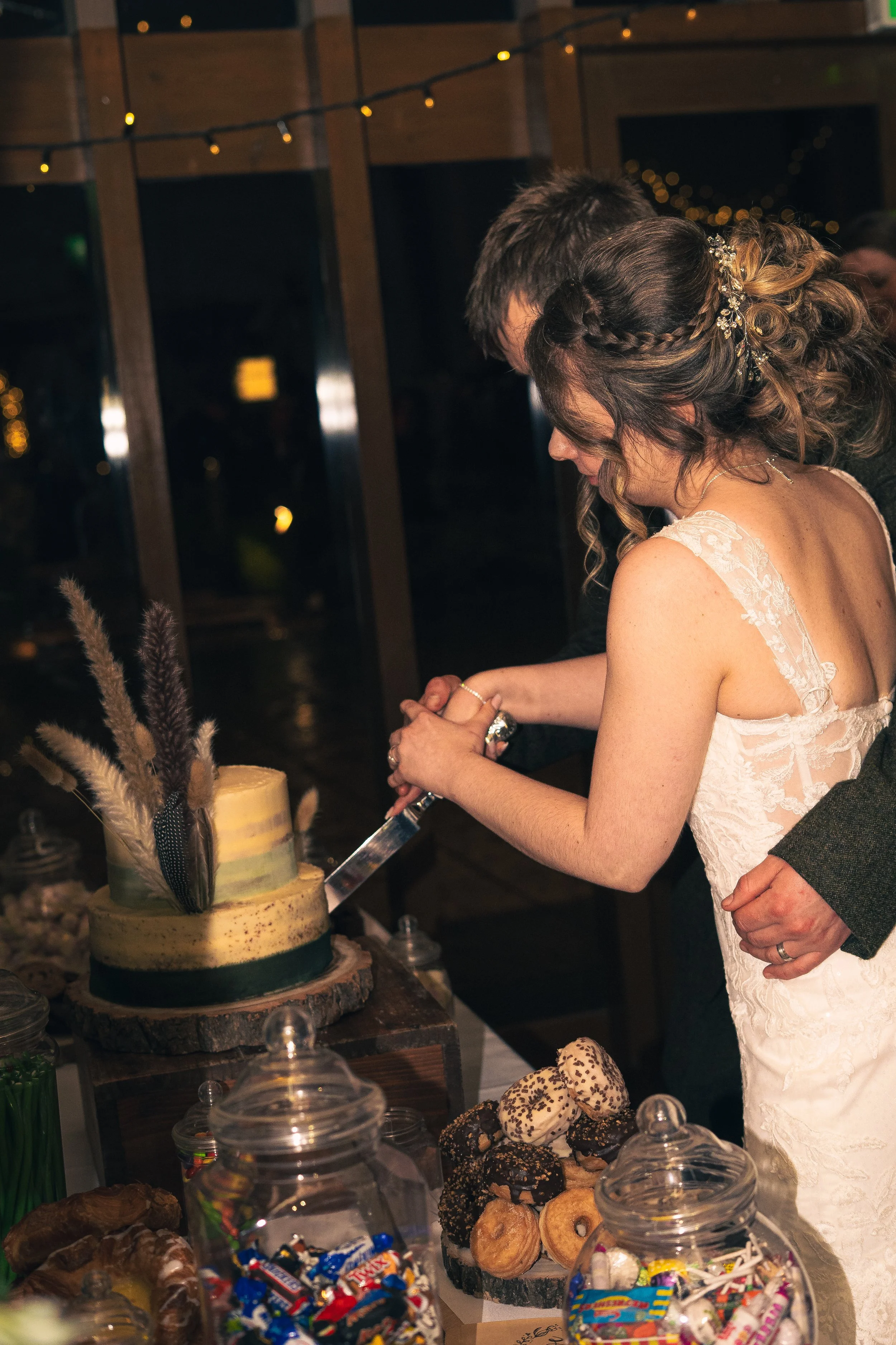 A bride and groom cut a wedding cake together at their celebration, with desserts and treats on a table nearby.