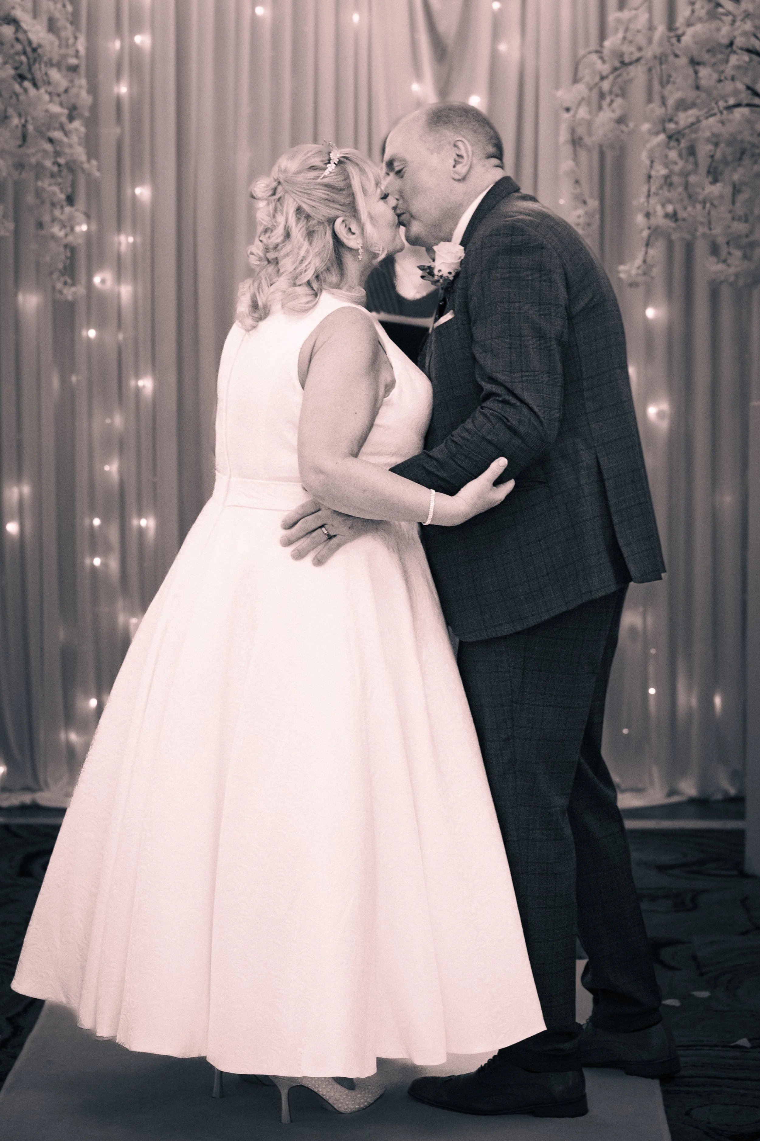 A couple, dressed in wedding attire, sharing a kiss during their wedding ceremony with a decorated backdrop of curtains and string lights.
