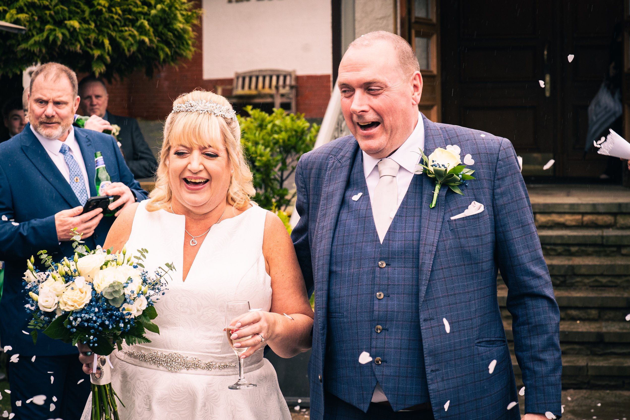 Bride and groom smiling and holding a glass of champagne, surrounded by wedding guests and confetti, outside a wedding venue.