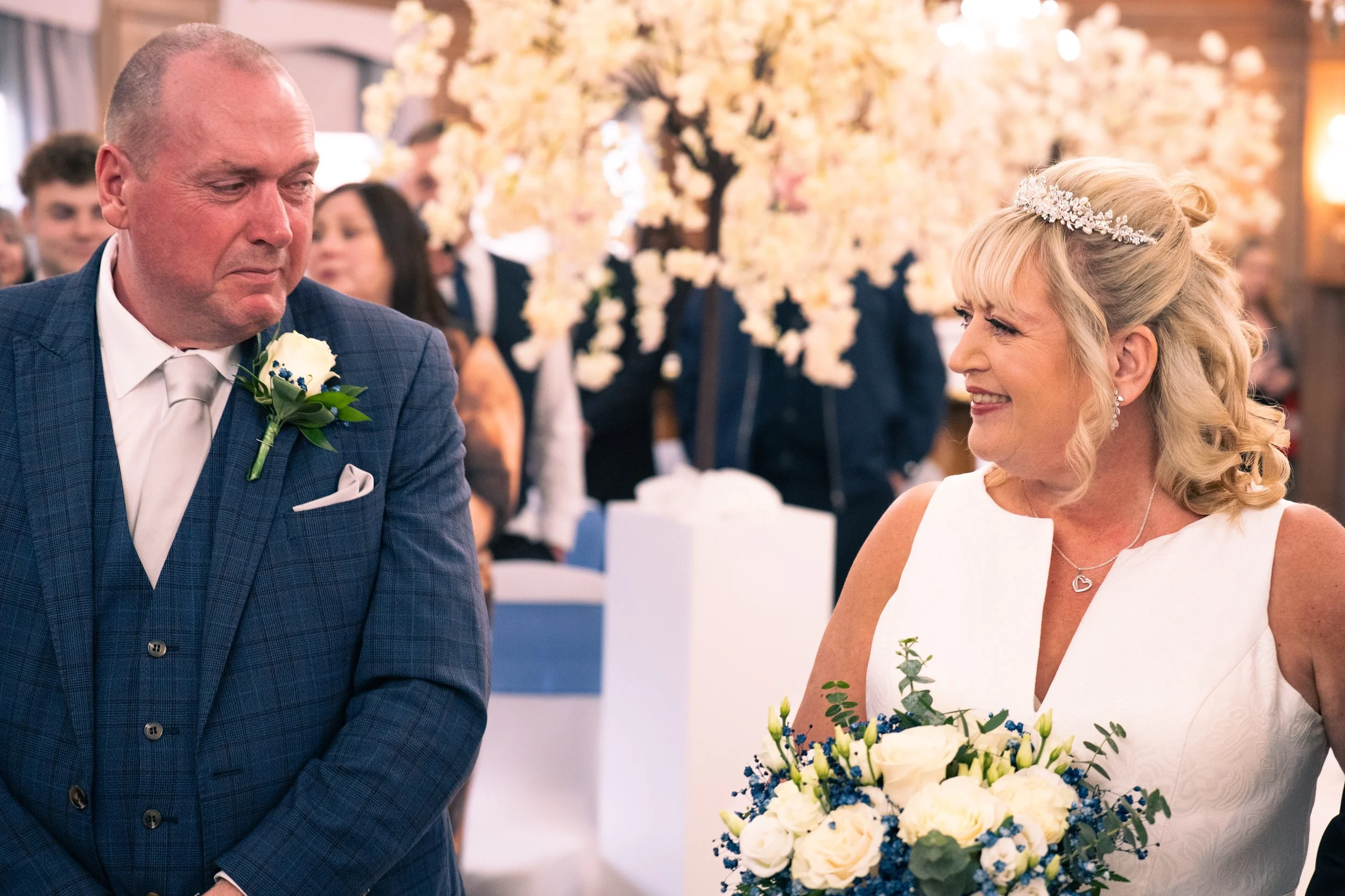 A bride and groom during a wedding ceremony. The bride is smiling and holding a bouquet of white roses and blue flowers. The groom is looking at her, wearing a blue checkered suit with a white shirt and a white tie. The background shows wedding guest