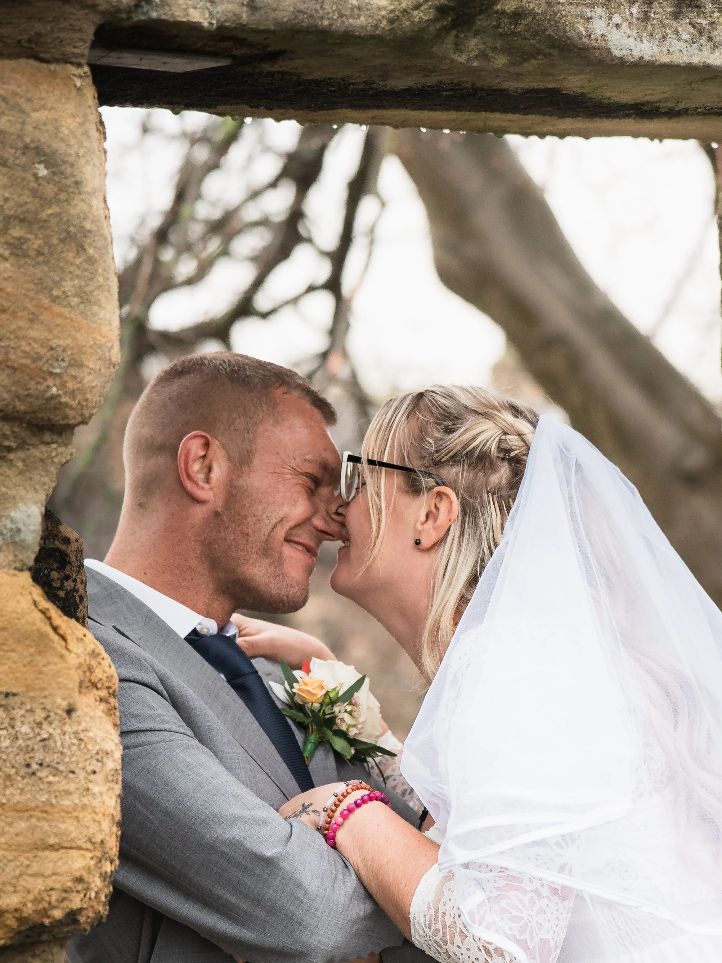 A couple, likely newlyweds, sharing a close, intimate moment outdoors, with their foreheads touching and eyes closed, under a natural archway formed by tree branches.