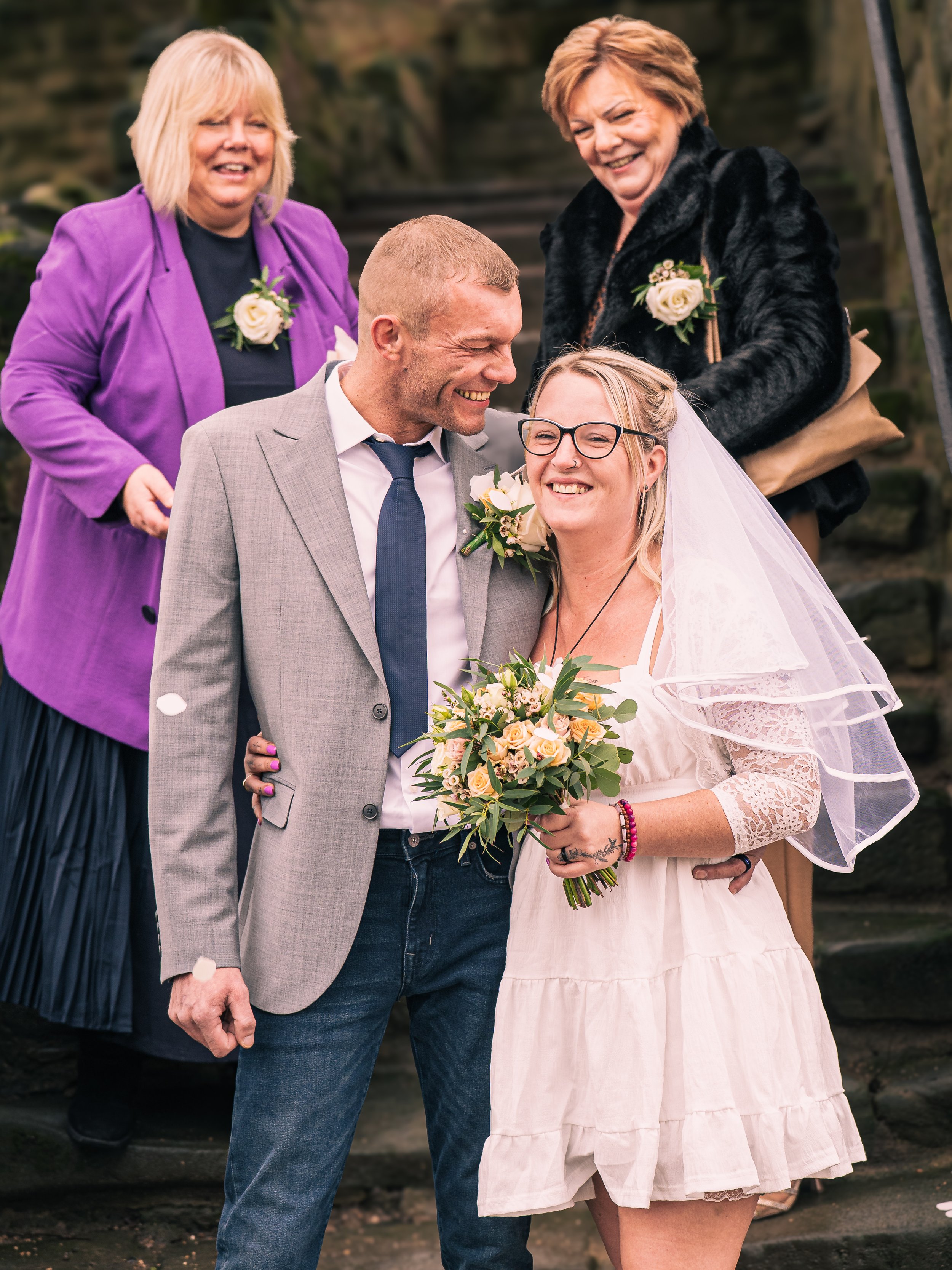 A bride and groom smiling and sharing a moment during their wedding ceremony, with two women in the background, all outdoors with stone steps behind them.