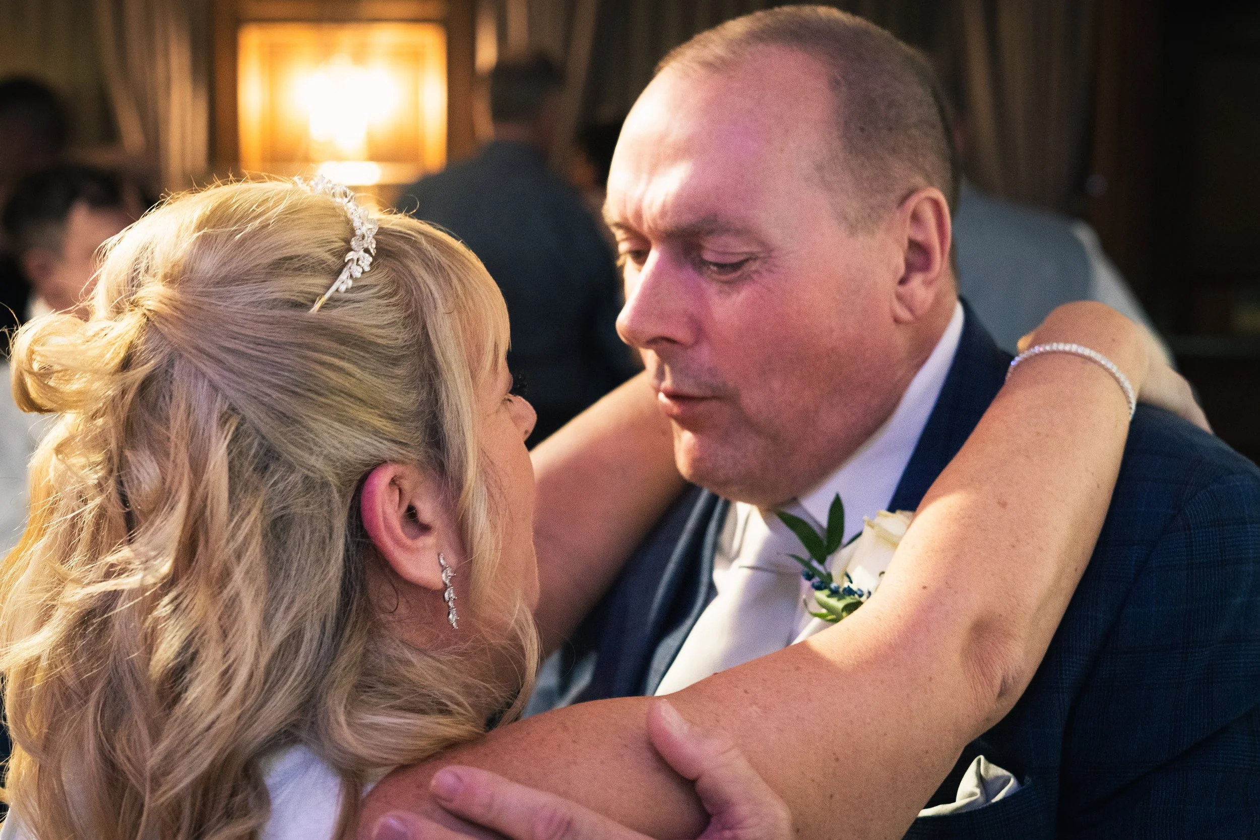 A bride and groom sharing a dance, gazing into each other's eyes, with emotional expressions. The bride has blonde, wavy hair with a decorative hairpiece and is wearing earrings. The groom has short hair, a boutonniere, and is dressed in a suit. The 