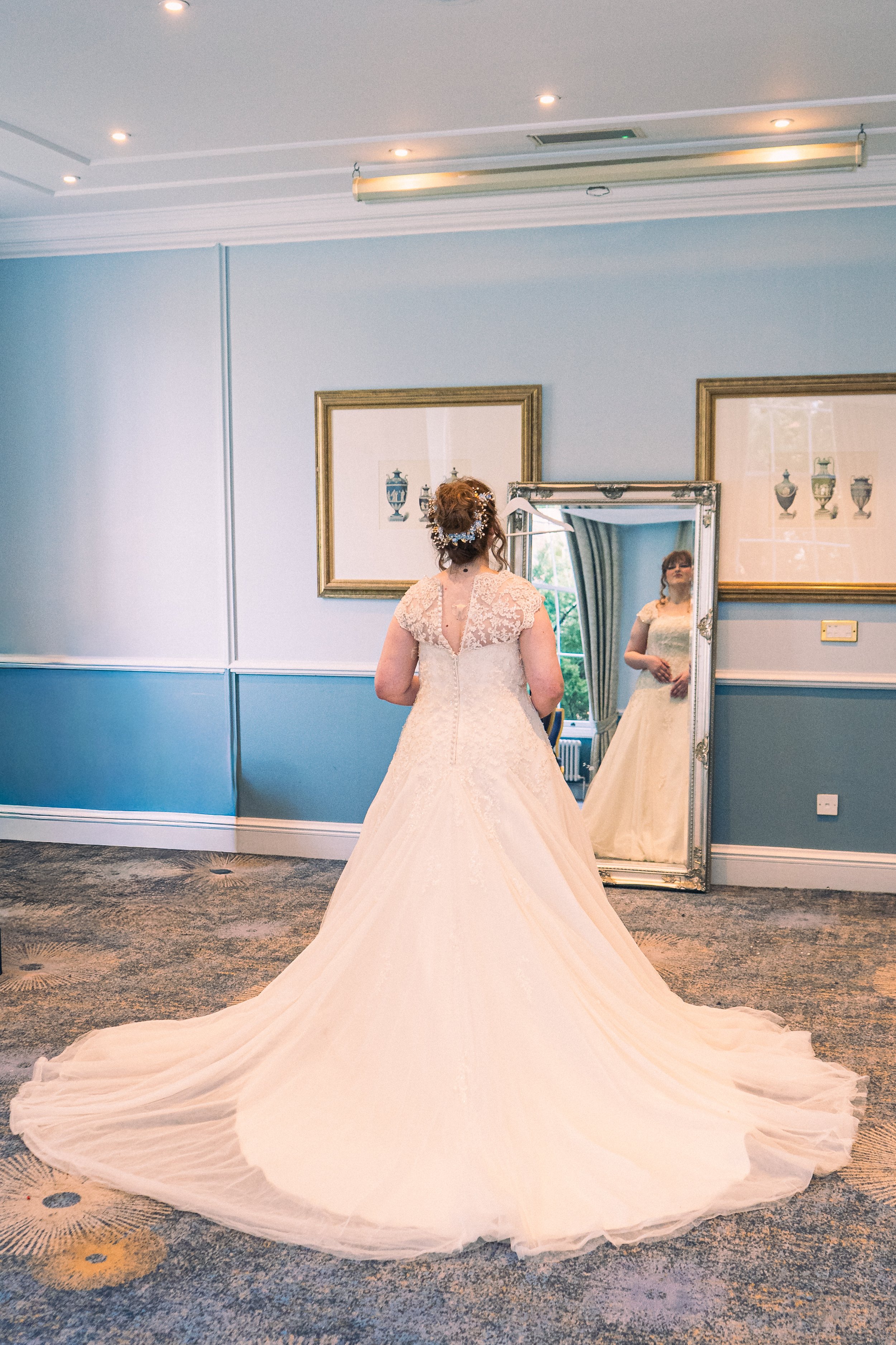 A bride in a wedding dress standing in front of a large mirror, looking at her reflection. The wedding dress has lace details and a long train, with her hair styled and adorned with a floral headpiece. The room has light blue walls with framed artwor