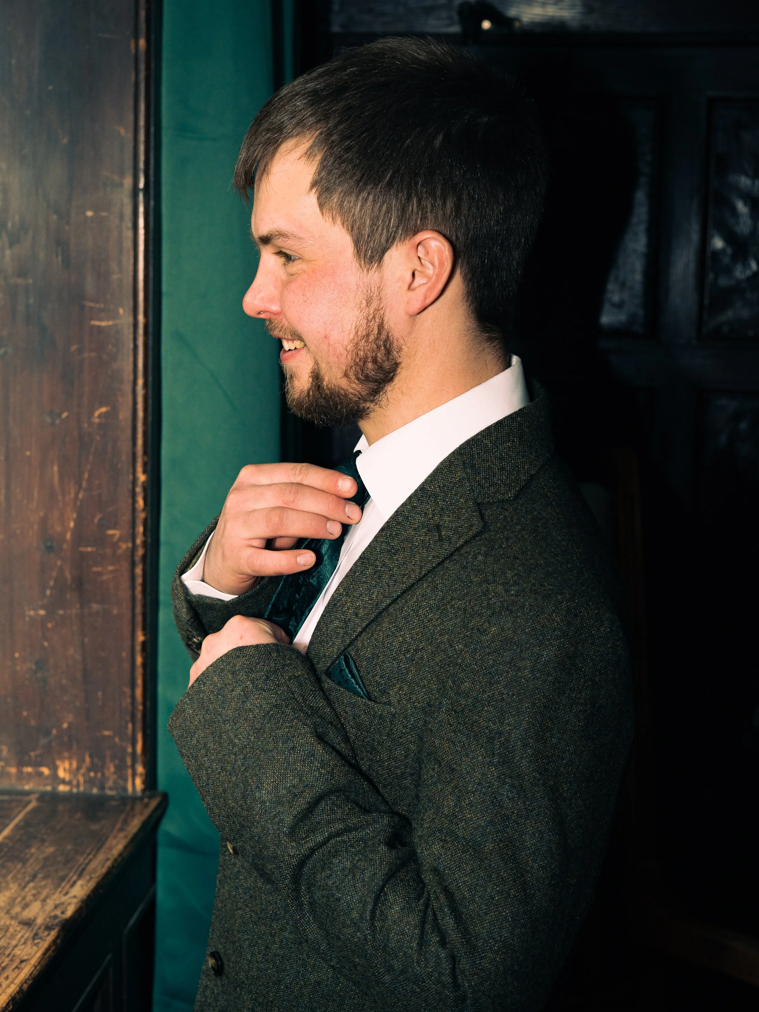 A man adjusting his tie while standing near a wooden surface, dressed in a formal suit with a white shirt and a dark jacket, smiling and looking towards the left.