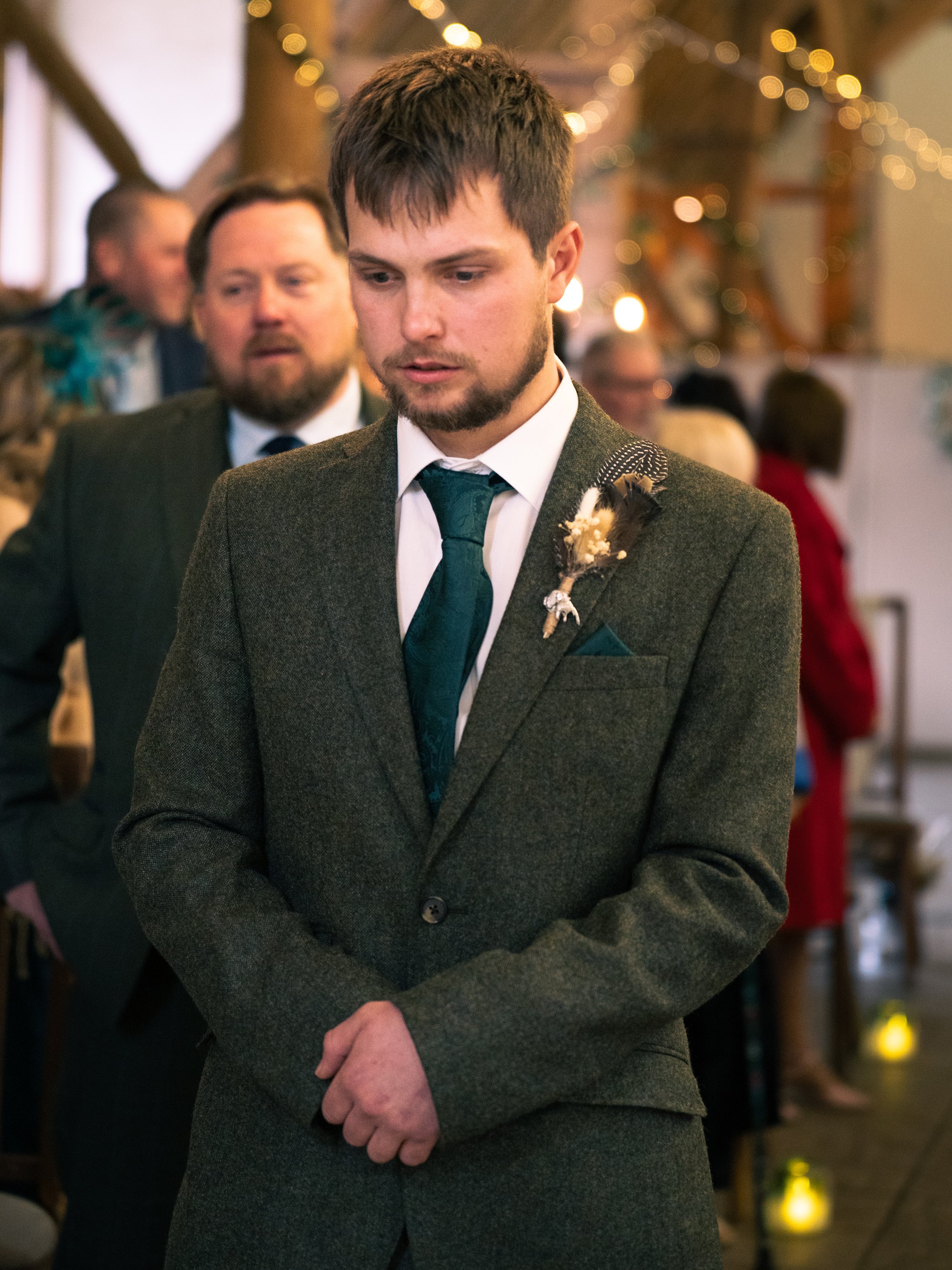 A young man in a gray suit with a dark green tie and boutonniere, standing in a room decorated for a wedding, with other guests in the background.