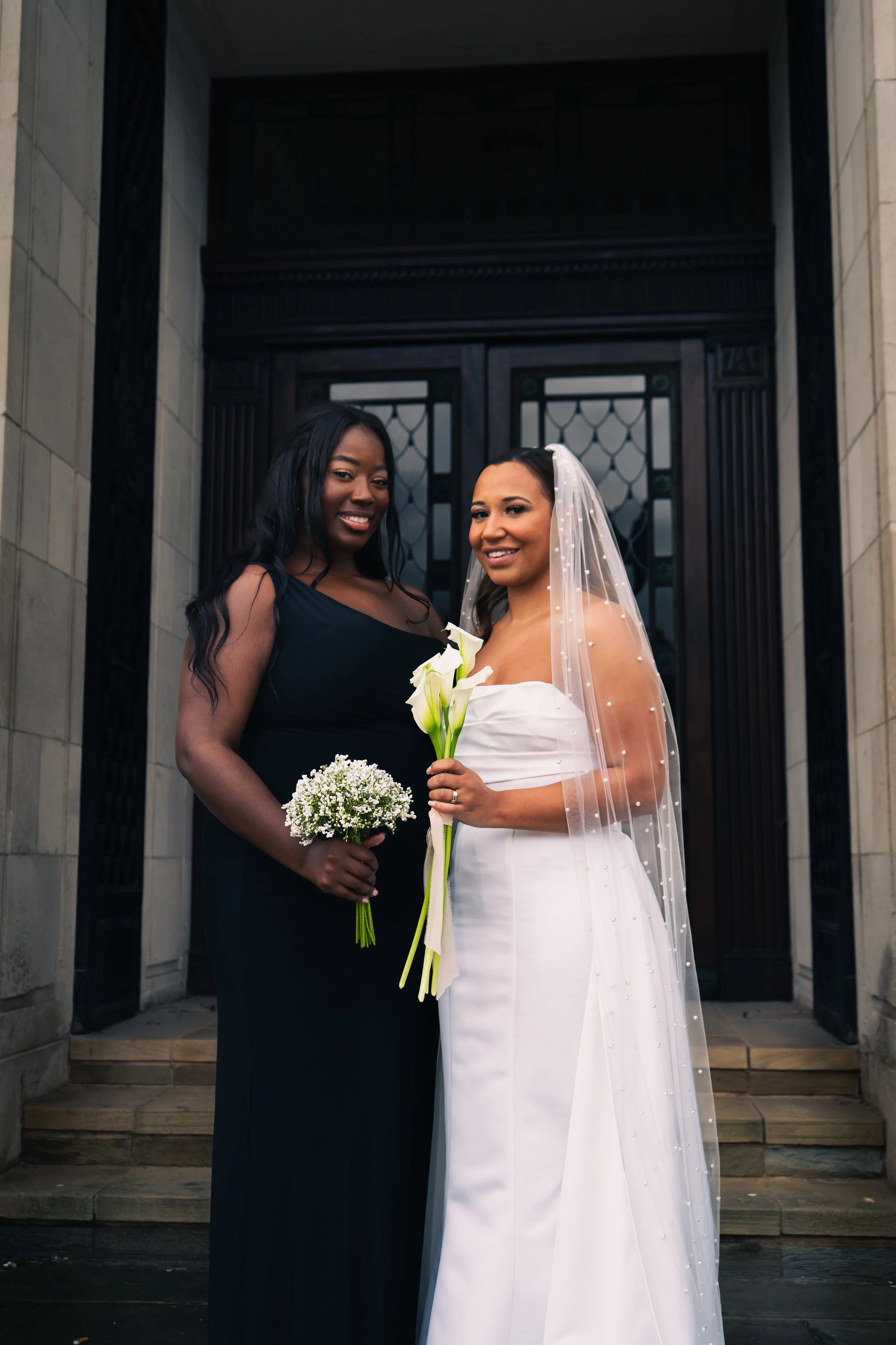 Two women standing on steps in front of a building, one in a white wedding dress with a veil, holding white calla lilies, and the other in a black dress holding white baby's breath, smiling at the camera.