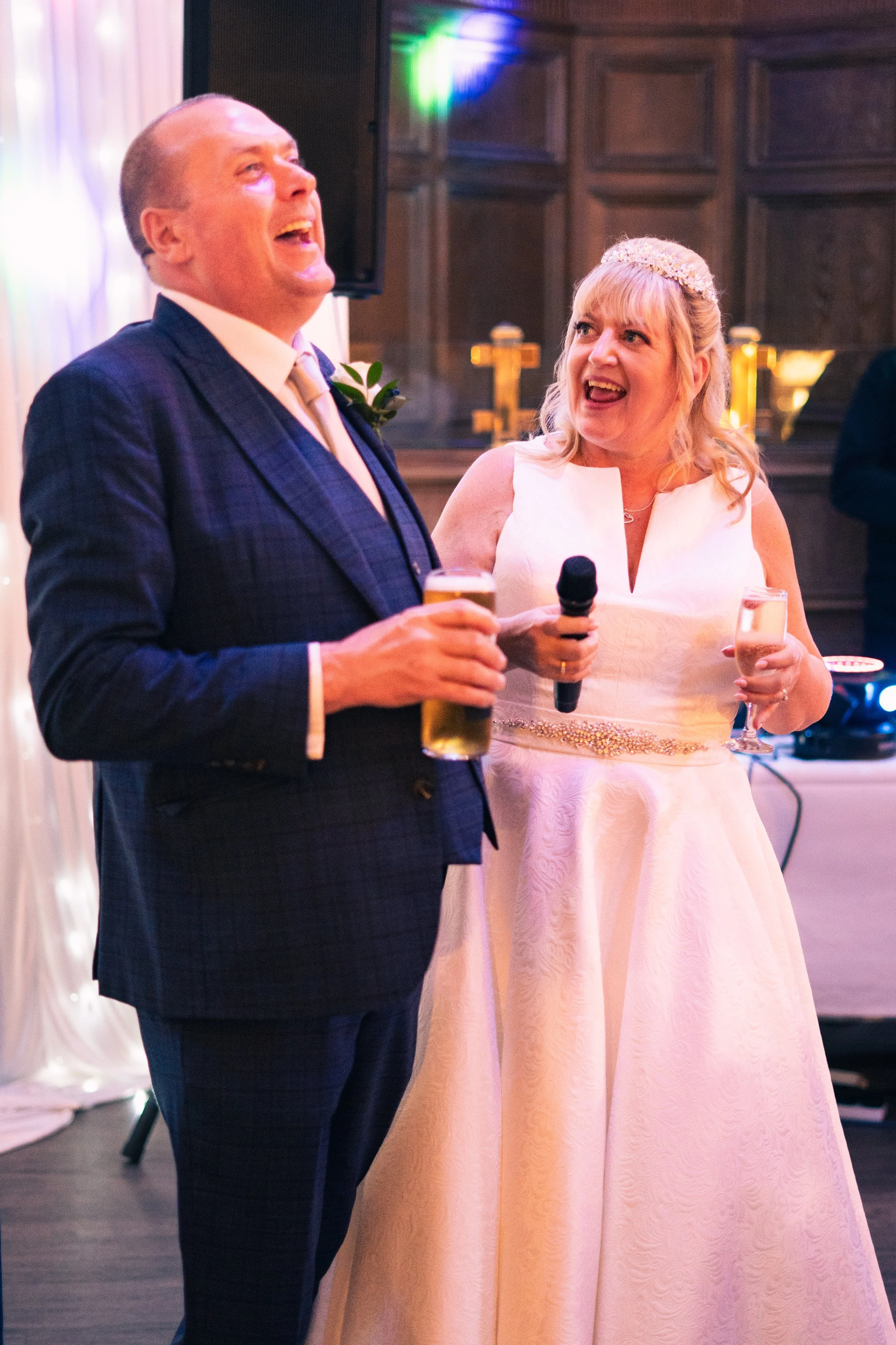 A man and woman at a wedding reception, standing side by side, smiling and laughing. The man is holding a glass of beer, and the woman is holding a microphone and a glass of champagne. The woman is wearing a white wedding dress with a beaded belt, an