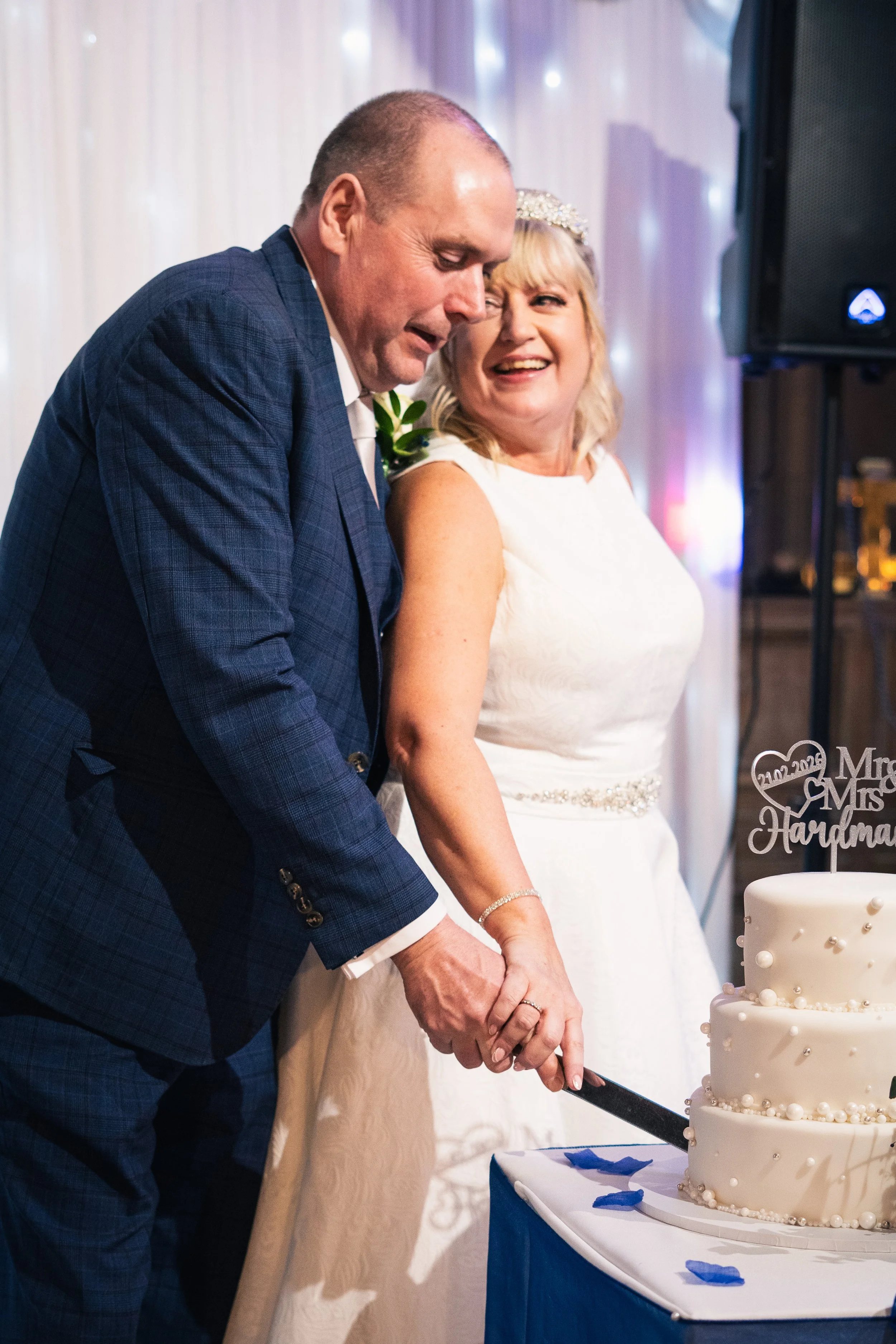 A bride and groom are cutting a three-tiered wedding cake together, holding a cake knife. The bride is smiling and wearing a white dress and headband, while the groom, in a blue plaid suit, looks down at the cake. There is a wedding topper on the cak
